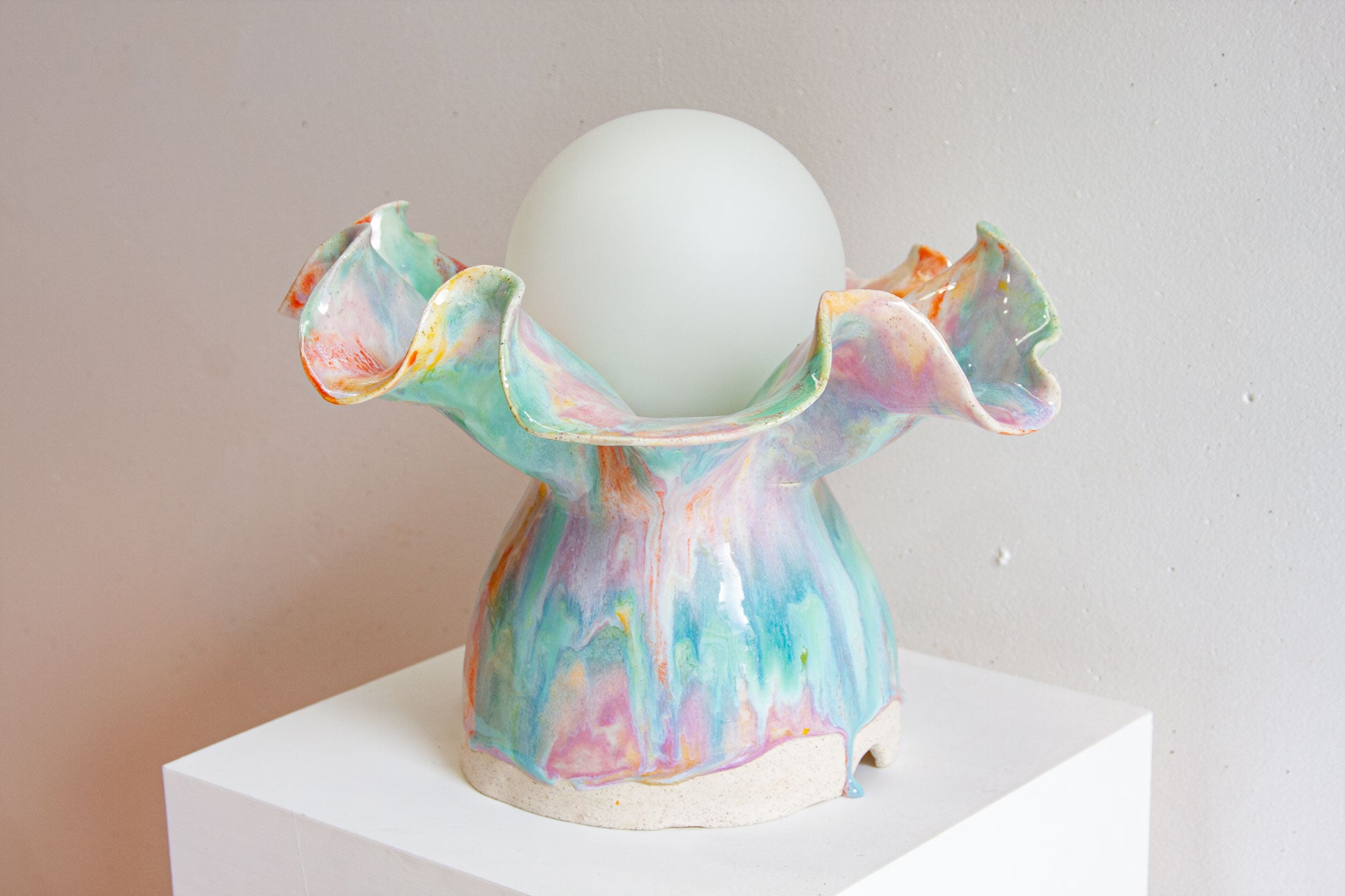 A handmade ceramic Oyster table lamp on a white plinth, the ruffled base glazed in purple, green, orange and yellow around a round white globe.
