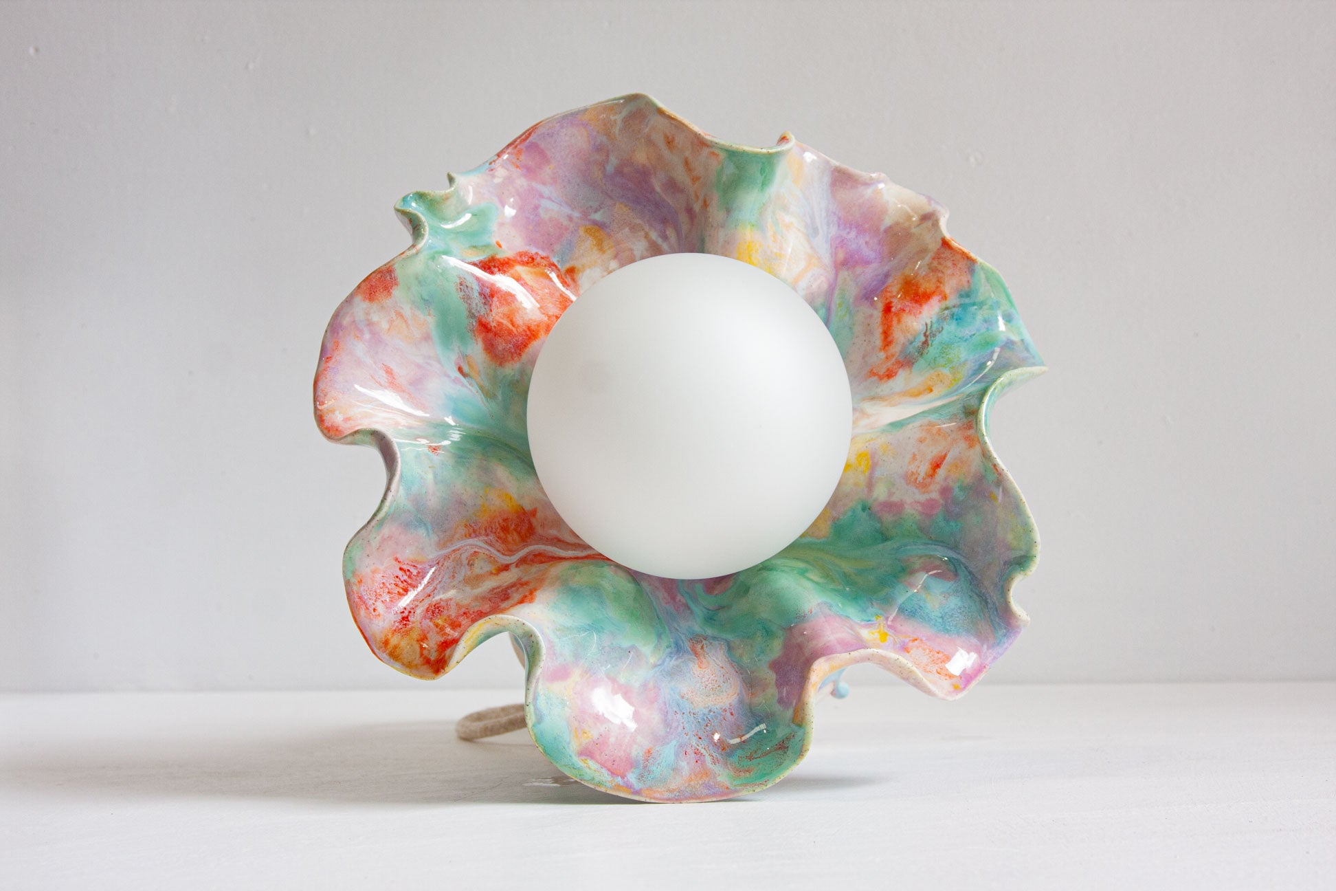 A handmade ceramic Oyster table lamp seen straight on, the wide ruffled rim in purple, green and orange surrounding the white globe.