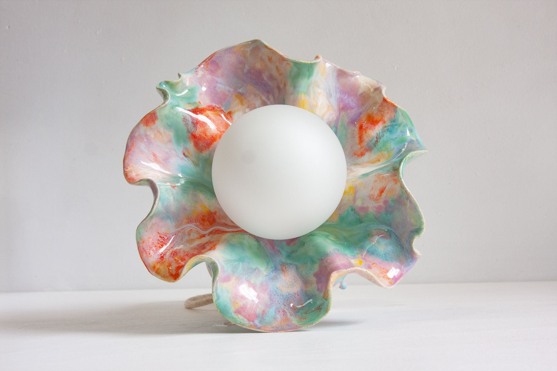 A handmade ceramic Oyster table lamp seen from the front, the wide ruffled rim glazed in purple, green and orange framing the lit globe.