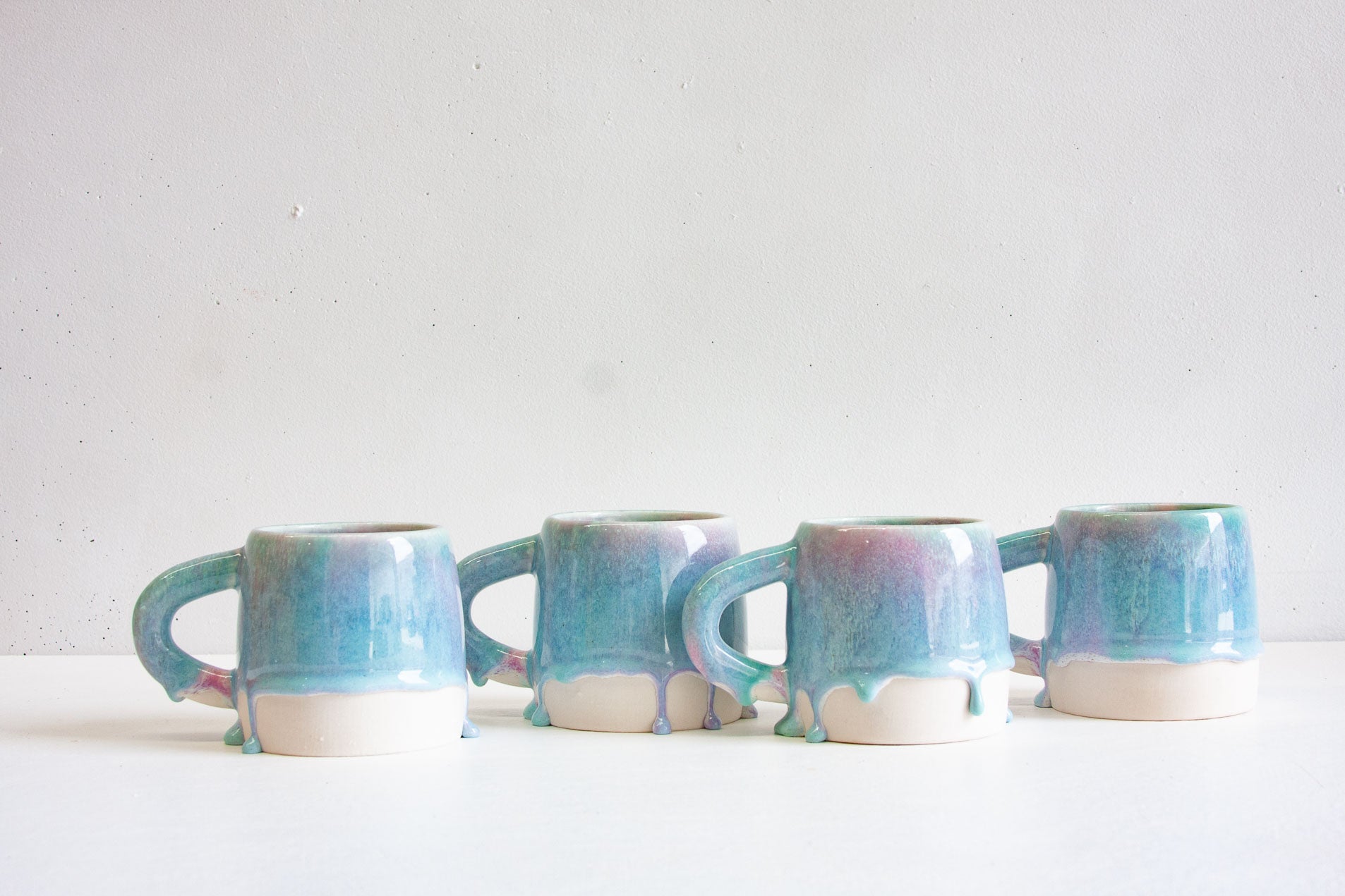 Four handmade ceramic large mugs in a row, the light purple and blue glaze running down into rounded drips over the bare white clay base.