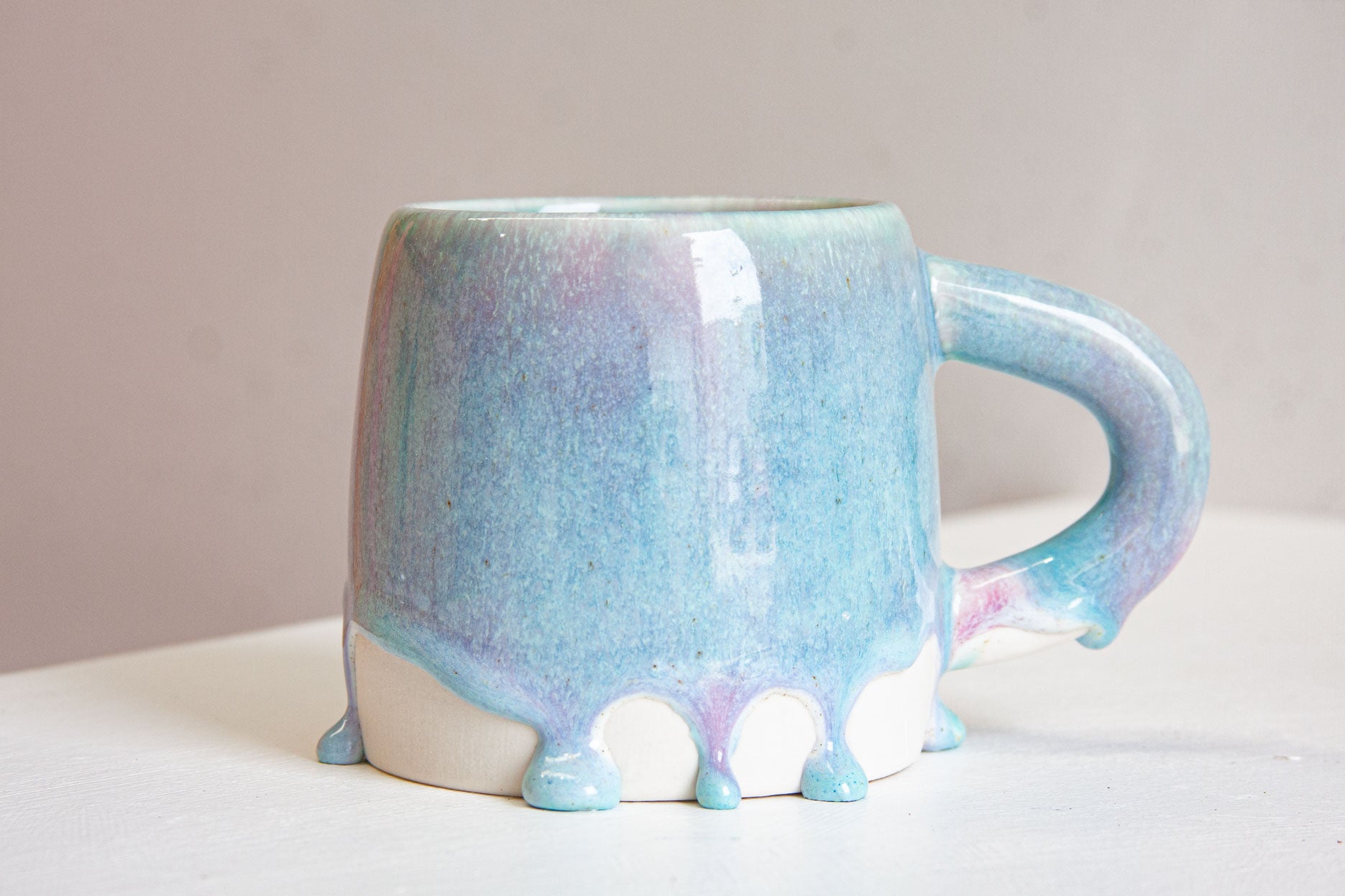 A handmade ceramic large mug, handle to the right, the light purple and blue glaze thinning at the rim and gathering into rounded drips above the bare white clay base.
