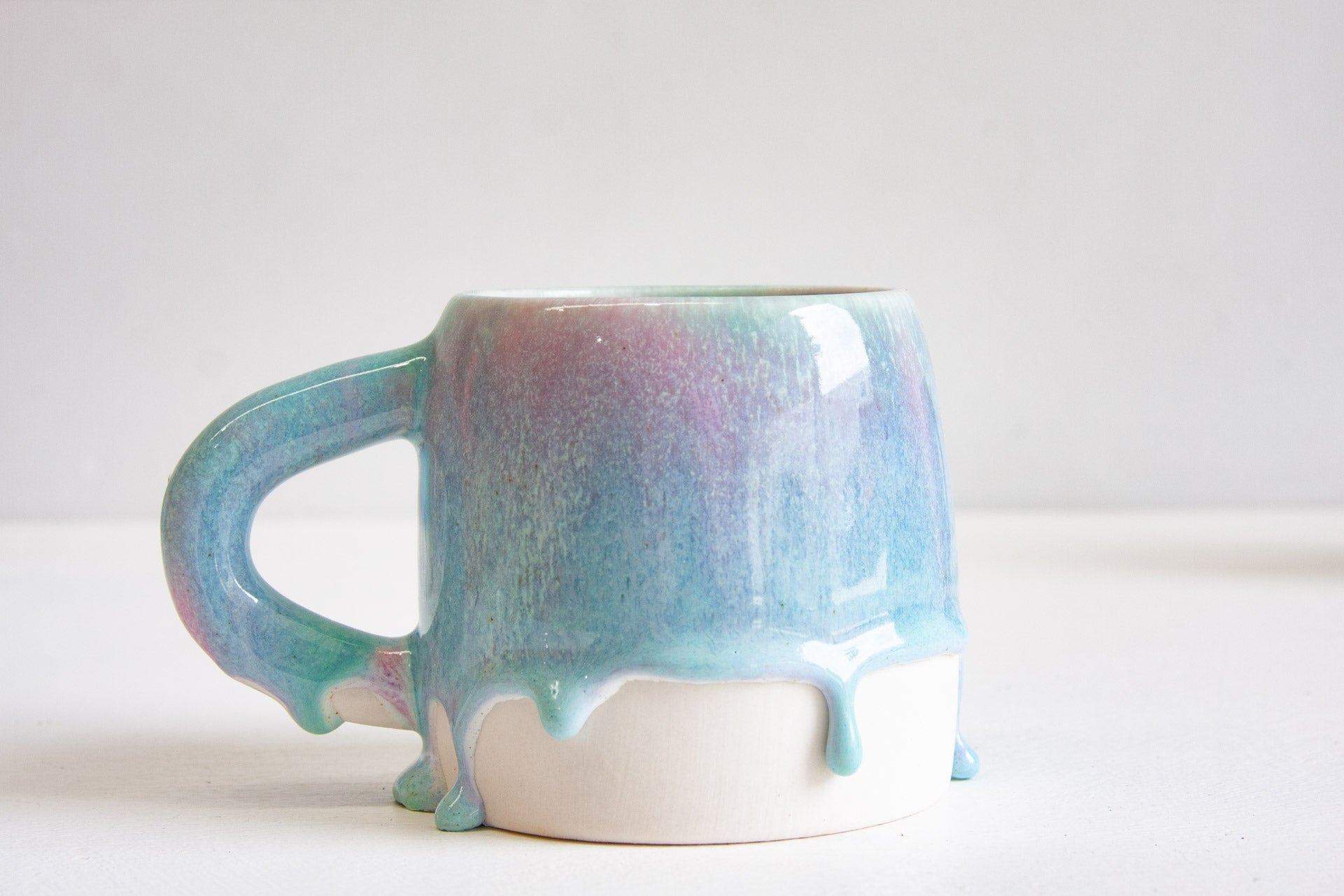 A handmade ceramic large mug, handle to the left, the light purple and blue glaze trailing down into drips over the bare white clay base.