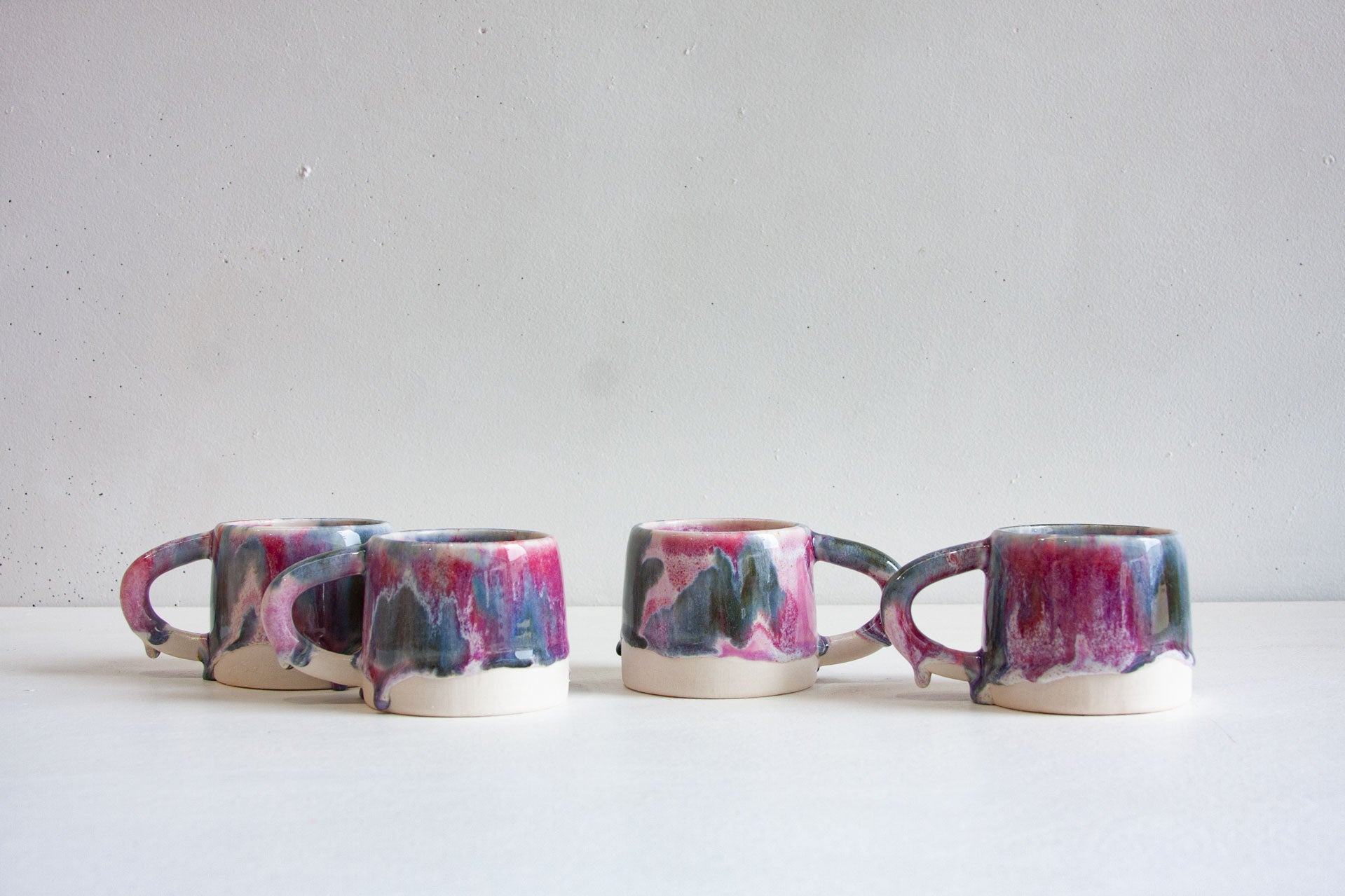 Four handmade ceramic mugs in a row, brush-glazed in light purple, dark purple and deep blue over white, with the glaze running to a bare clay base.