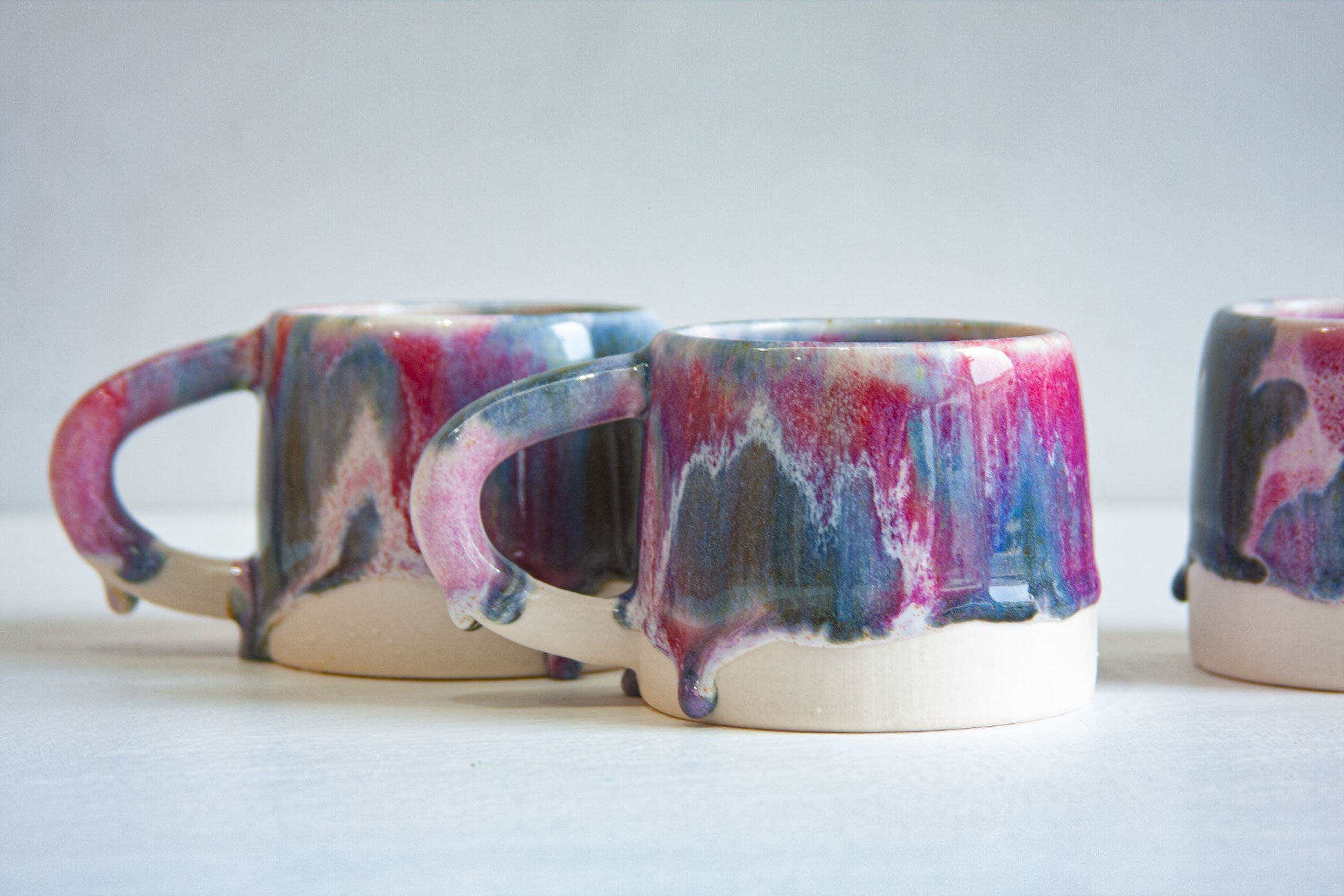Three handmade ceramic mugs close up, the purple and deep blue glaze breaking into white where it thins above the bare clay base.