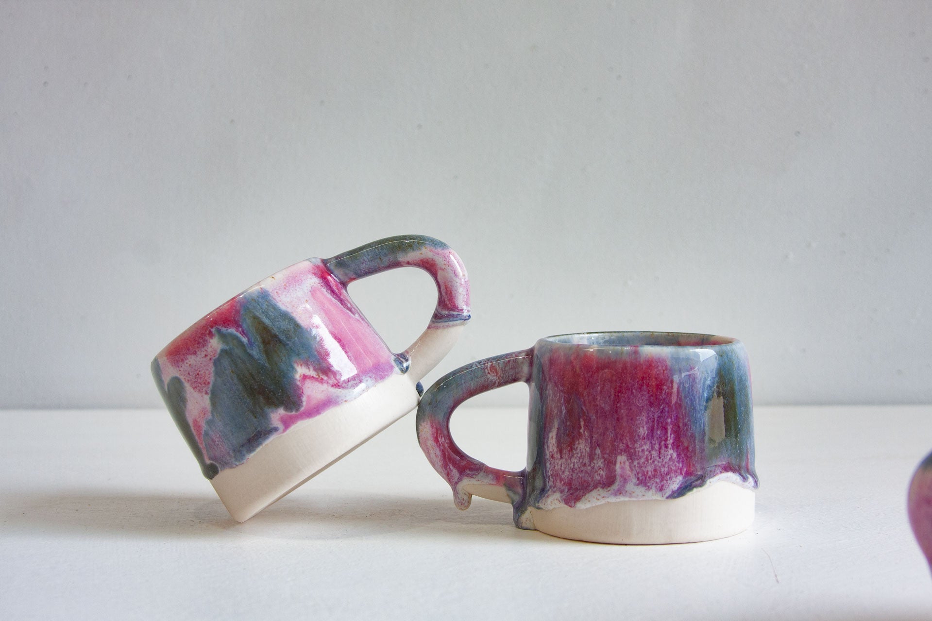 Two handmade ceramic mugs, one tipped on its side, both glazed in light and dark purple with deep blue running down to a bare white clay base.
