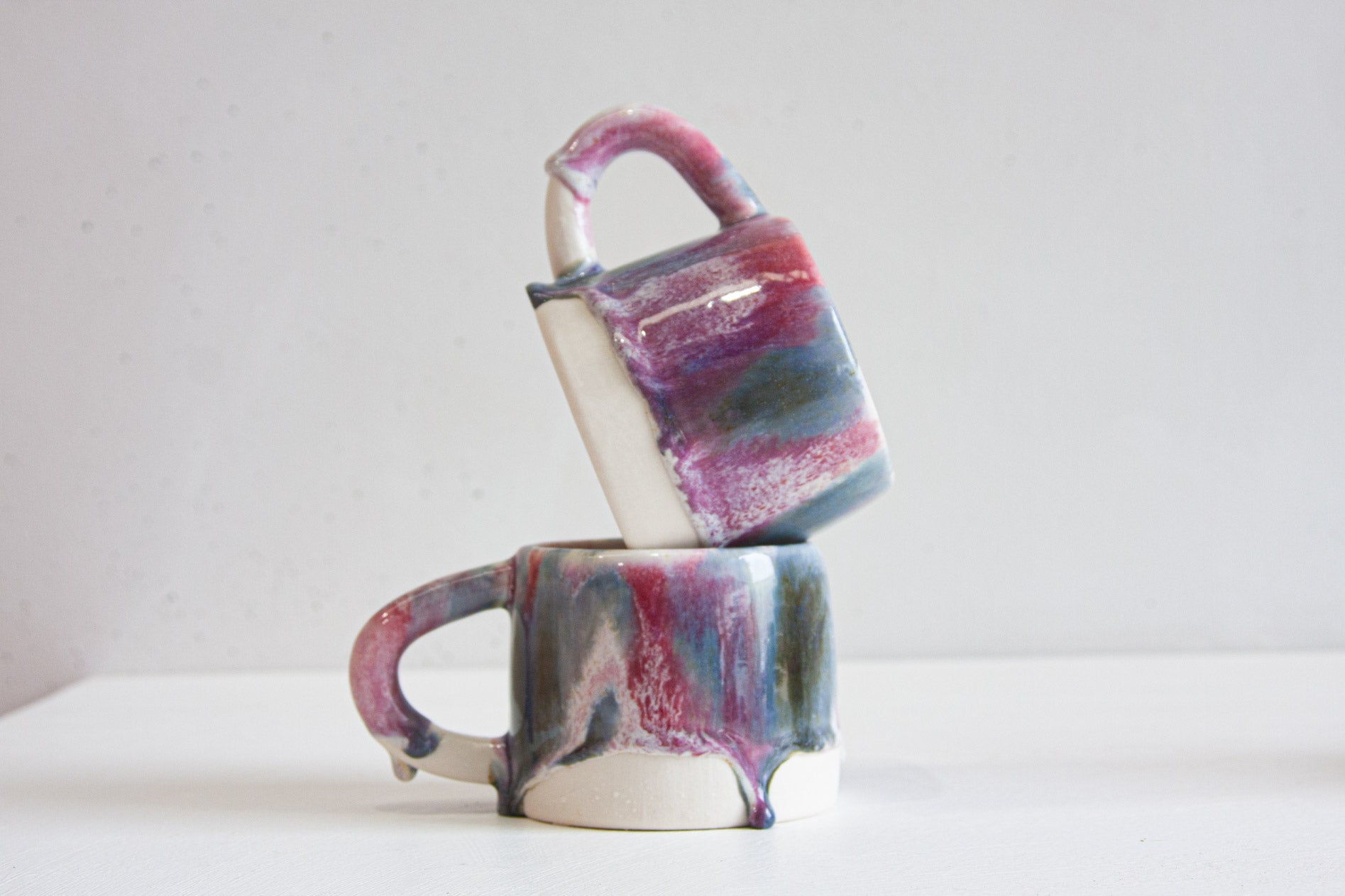 Two handmade ceramic mugs stacked, one tilted on top of the other, showing the purple and blue glaze running over the handles.