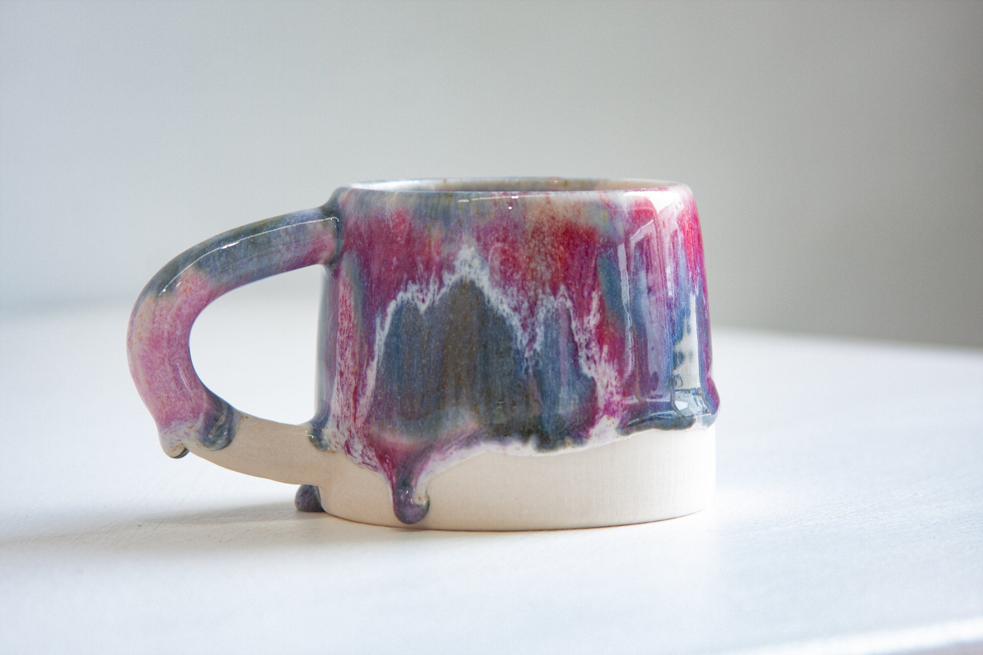 A single handmade ceramic mug seen side on, the light purple, dark purple and deep blue glaze dripping over a bare white clay base.