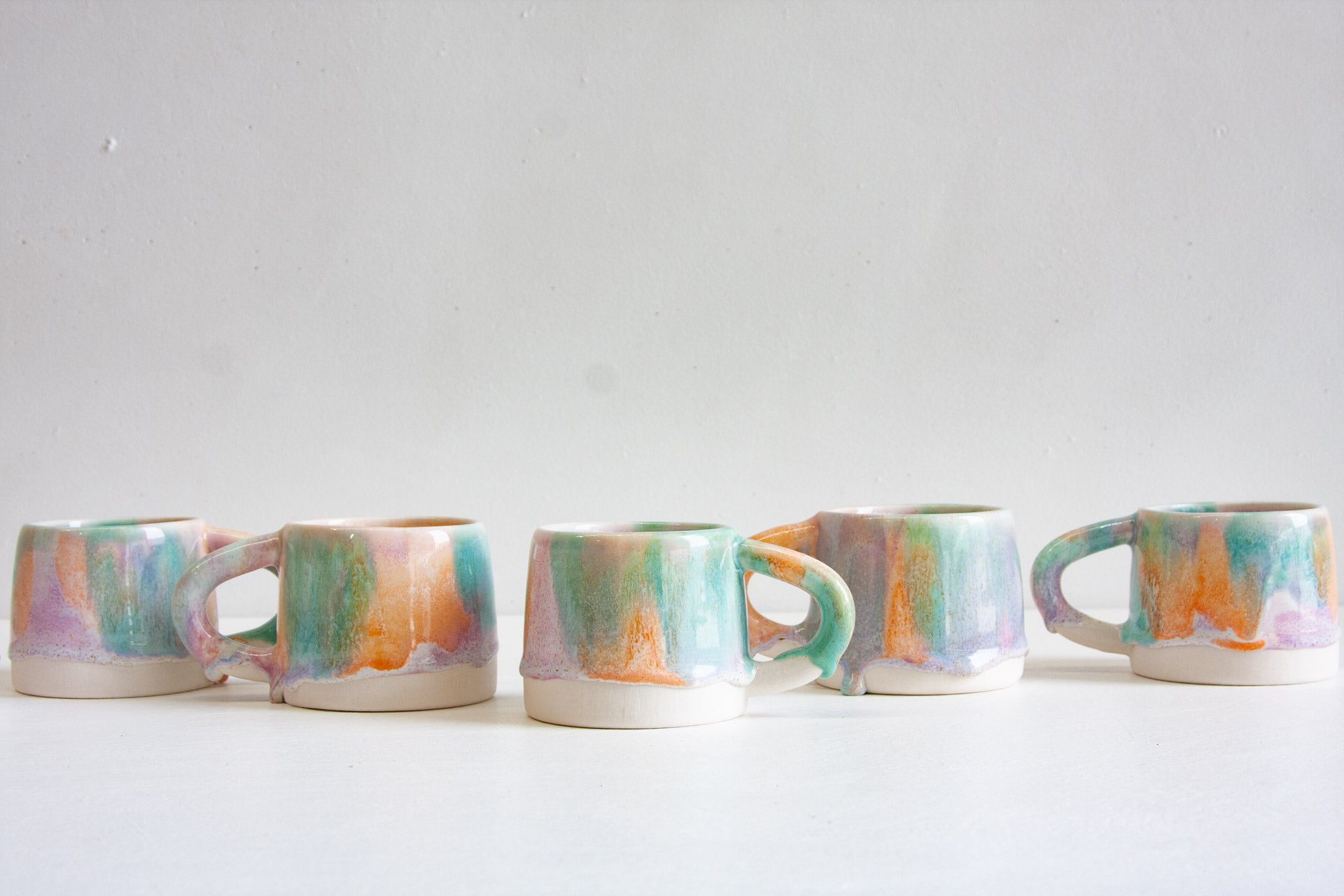 Five handmade ceramic mugs in a row, each brush-glazed in pastel purple, green and orange over a bare white clay base.