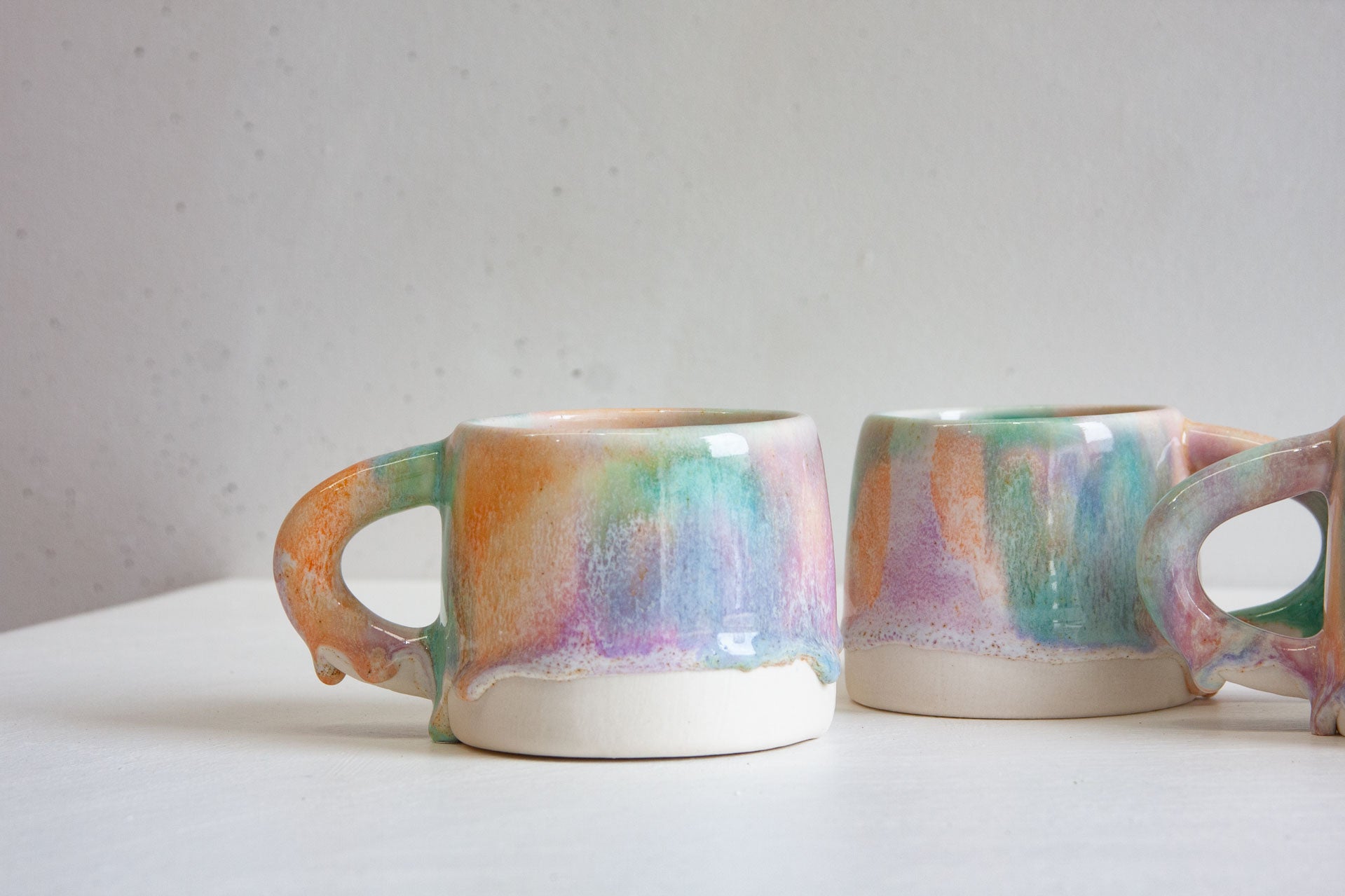 Two handmade ceramic mugs side by side, the pastel purple, green and orange glaze running down into a soft line over the bare white clay base.