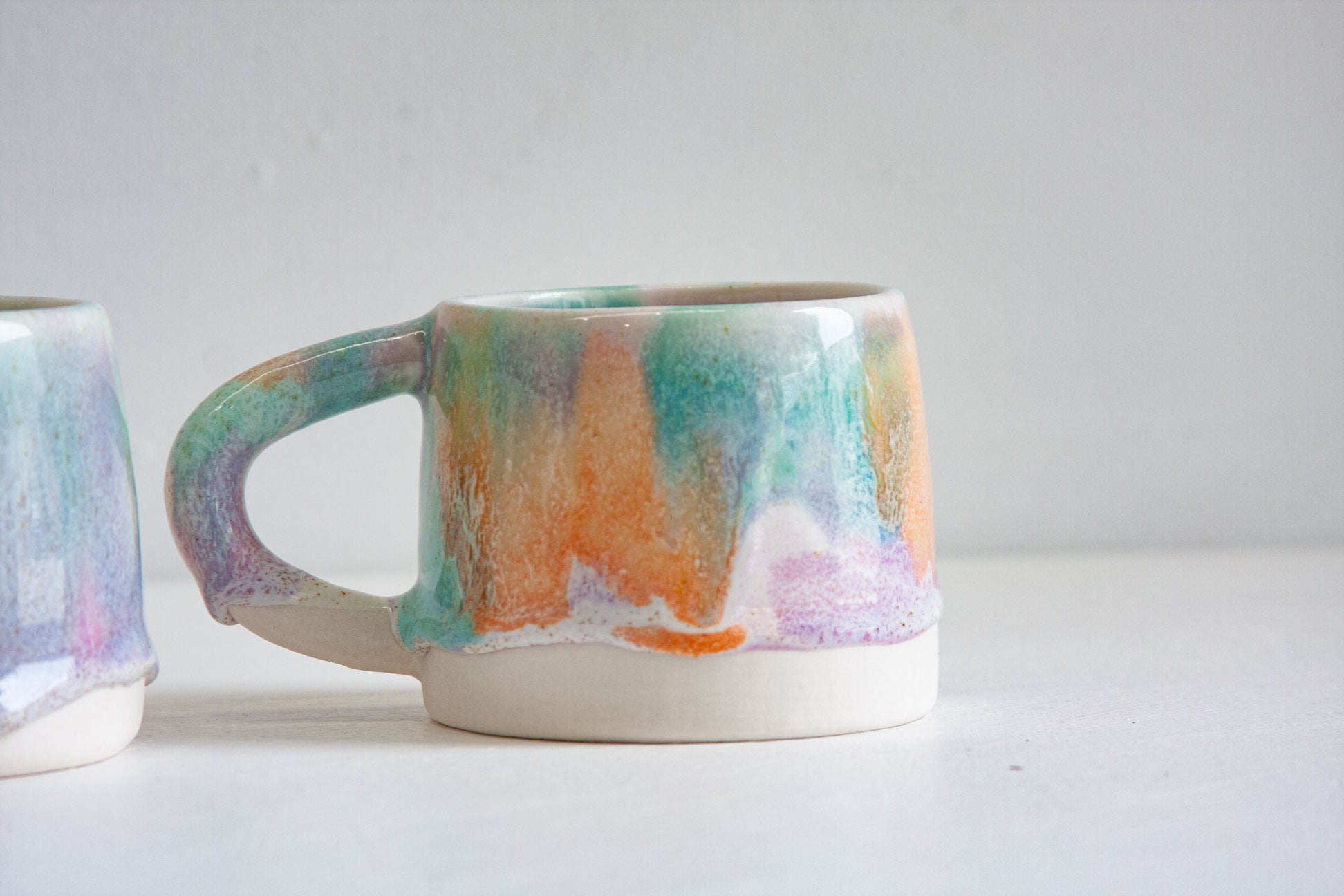 Close-up of a handmade ceramic mug, the green, orange and purple glaze running in soft vertical bands down to the bare white clay base.