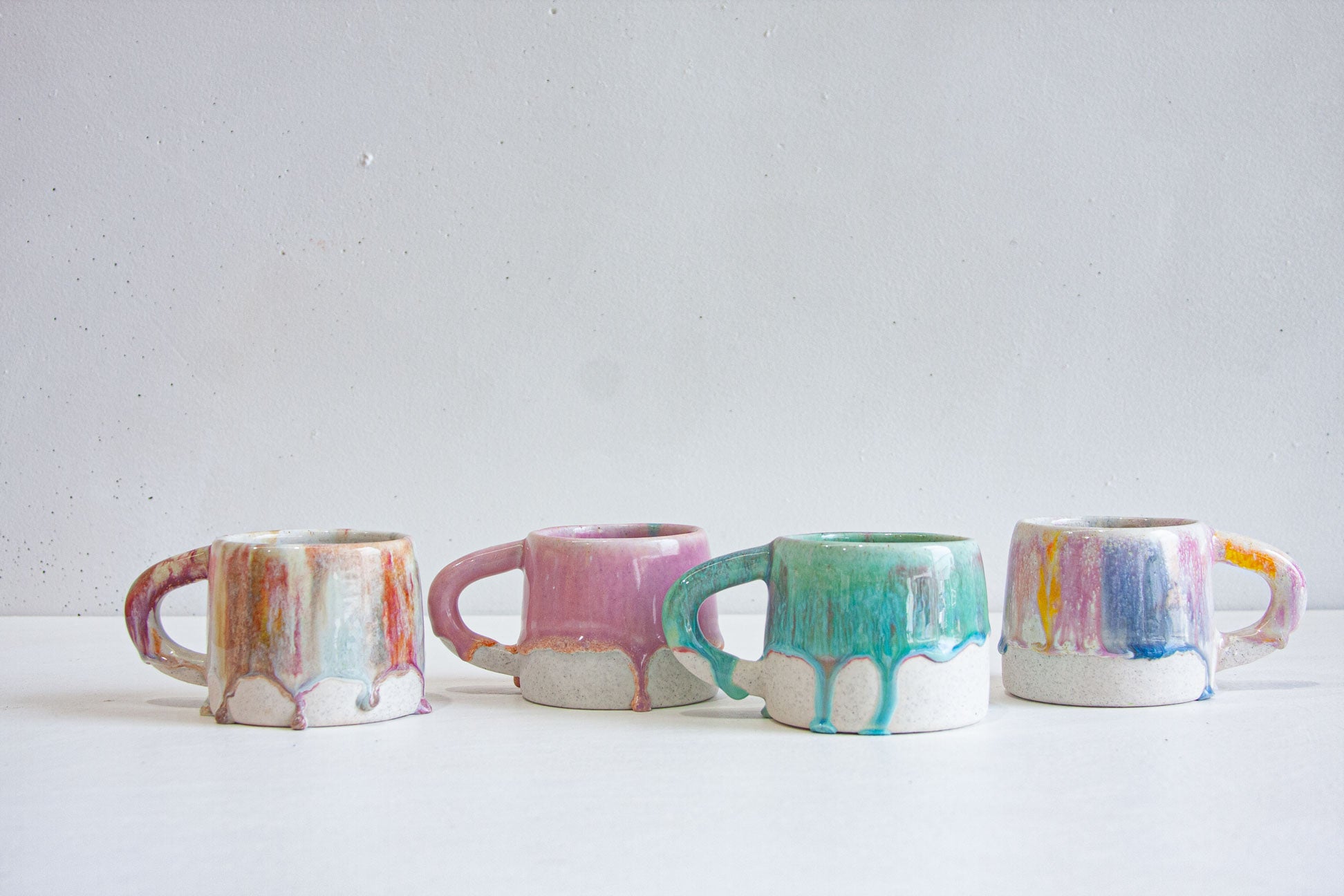 Four handmade ceramic mugs in a row: a pale cream streaked with peach and blue, a soft pink, a green and teal, and a speckled white run through with blue and yellow.
