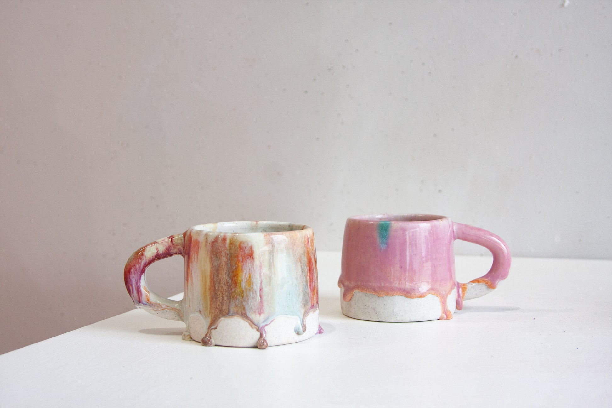 Two handmade ceramic mugs, one a pale cream streaked with peach, rust and blue, the other a soft pink with peach drips gathering above the bare clay base.