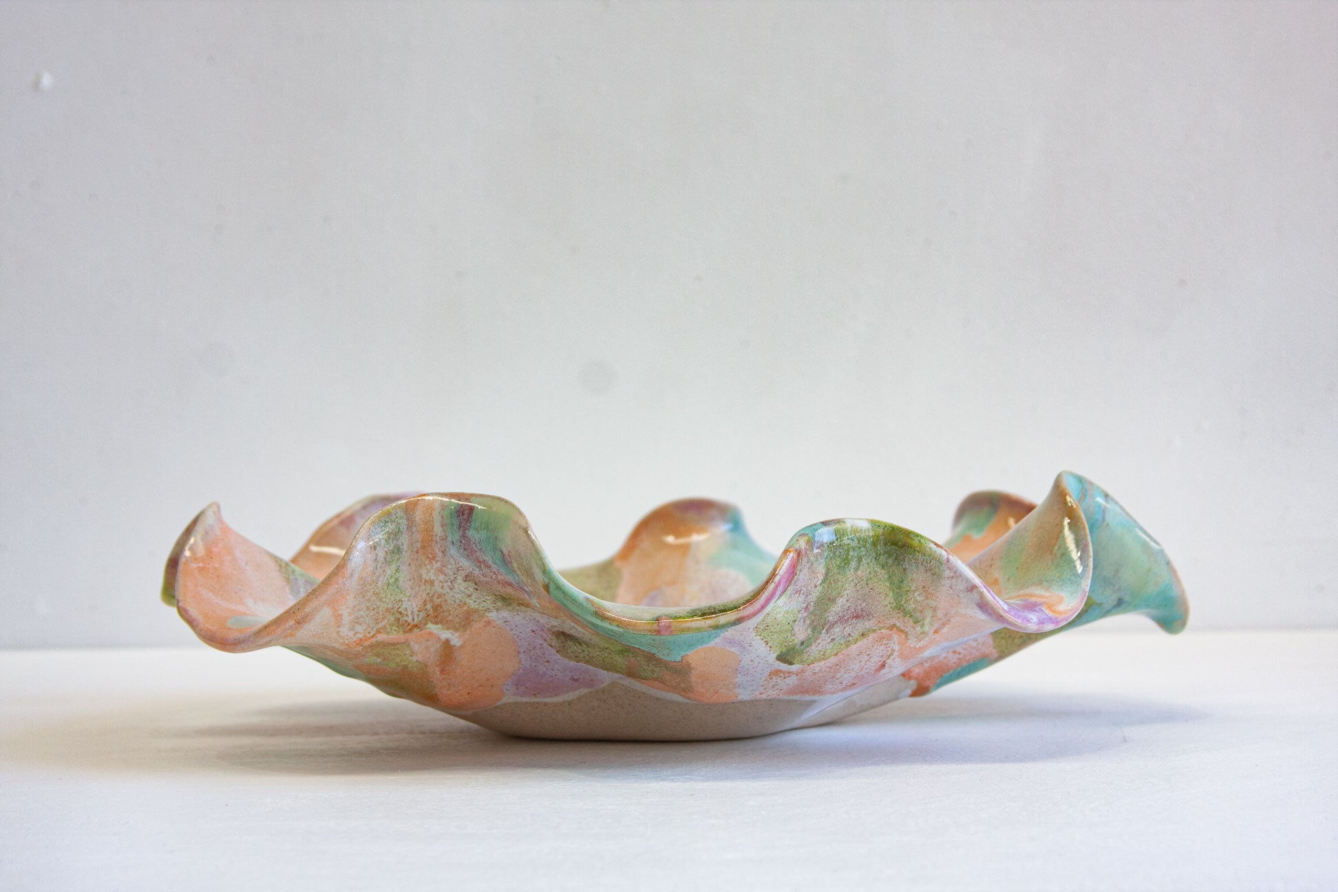 A handmade ceramic oyster platter seen side on, the low ruffled form showing peach and green across the rim above the bare clay foot.