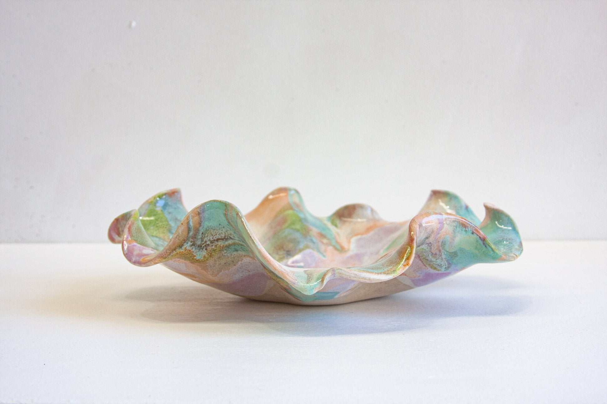 A handmade ceramic oyster platter seen from the side, its ruffled rim rising in waves, glazed in purple, green, peach and white.