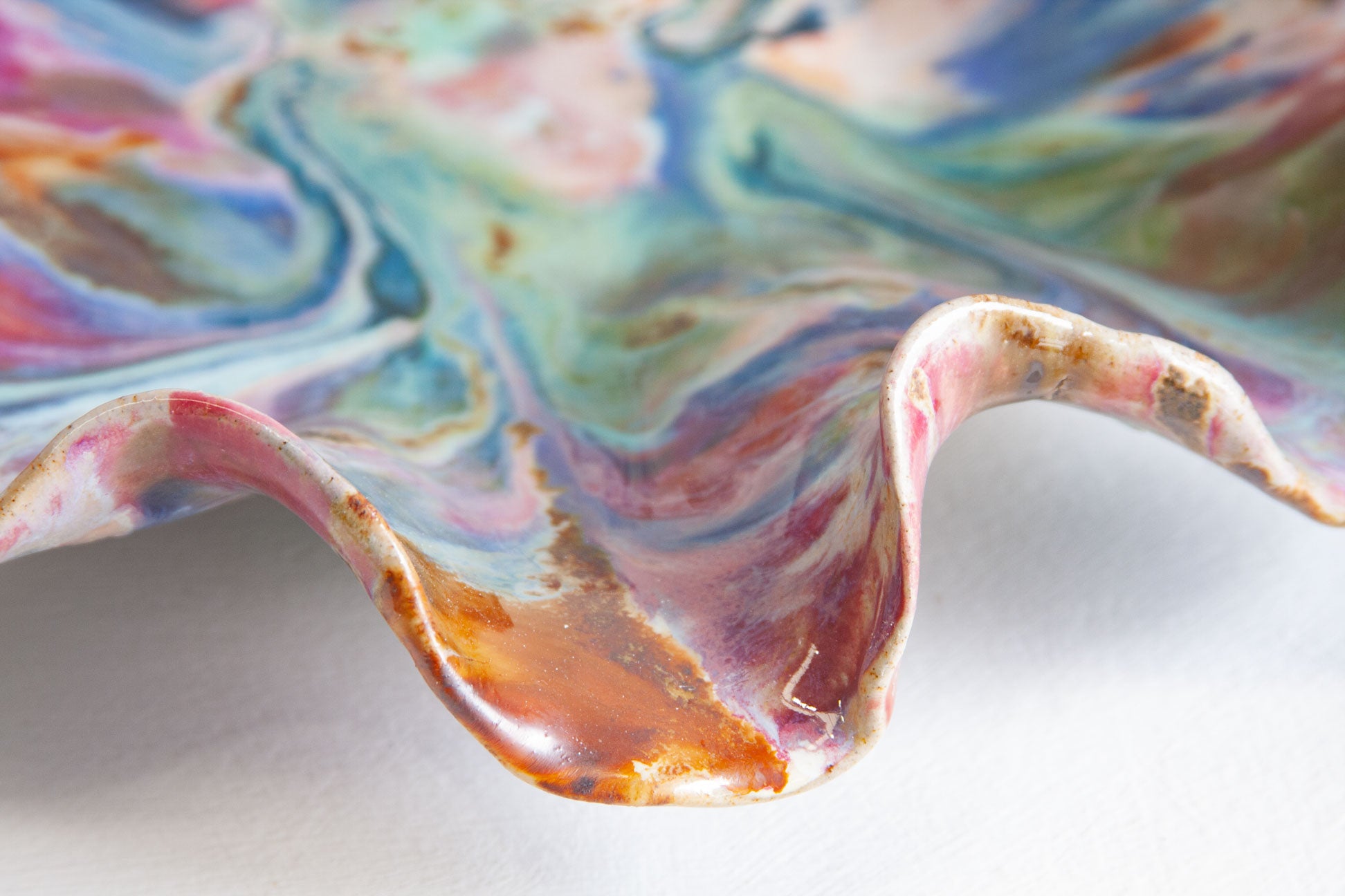 Close-up of one fold of a handmade ceramic oyster platter, the glaze running through rust brown, pink and blue along the curved edge.