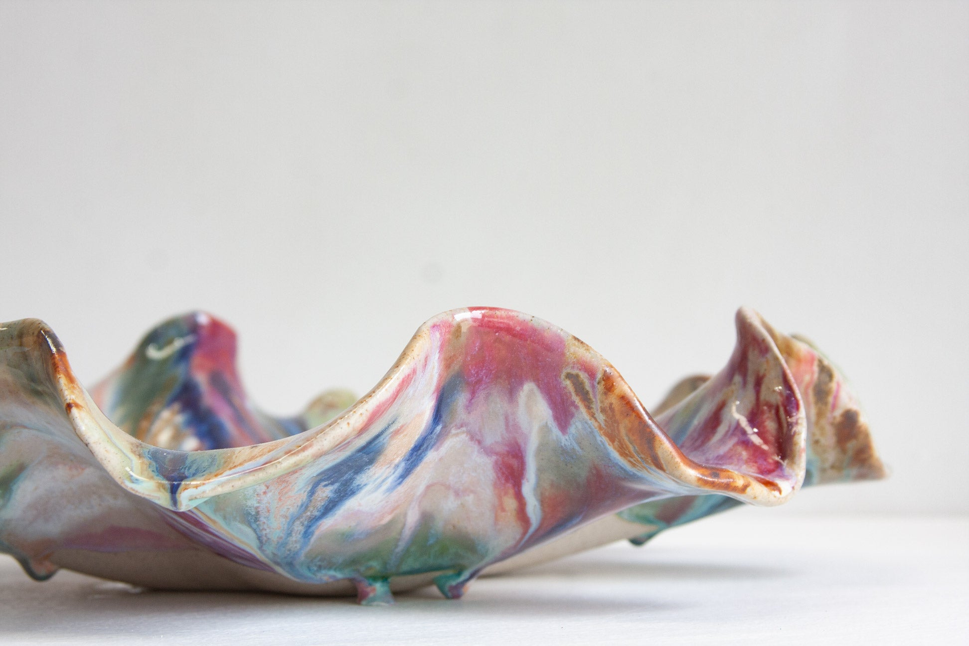 A handmade ceramic oyster platter seen from the side at table height, its wavy folds marbled in pink, blue, green and rust brown.
