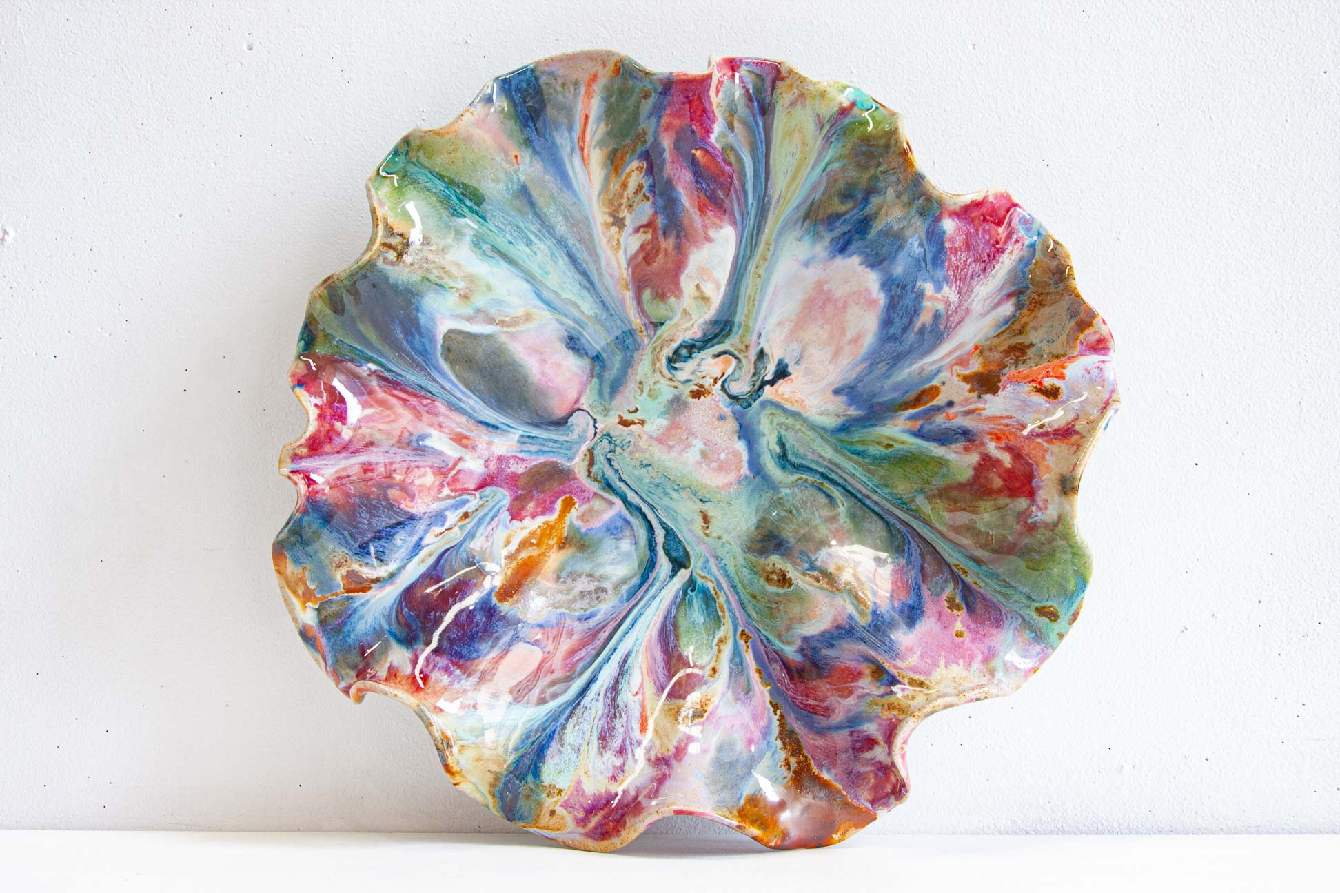 A handmade ceramic oyster platter seen from above, the ruffled flower-like rim glazed in green, pink, peach, brown and blue.