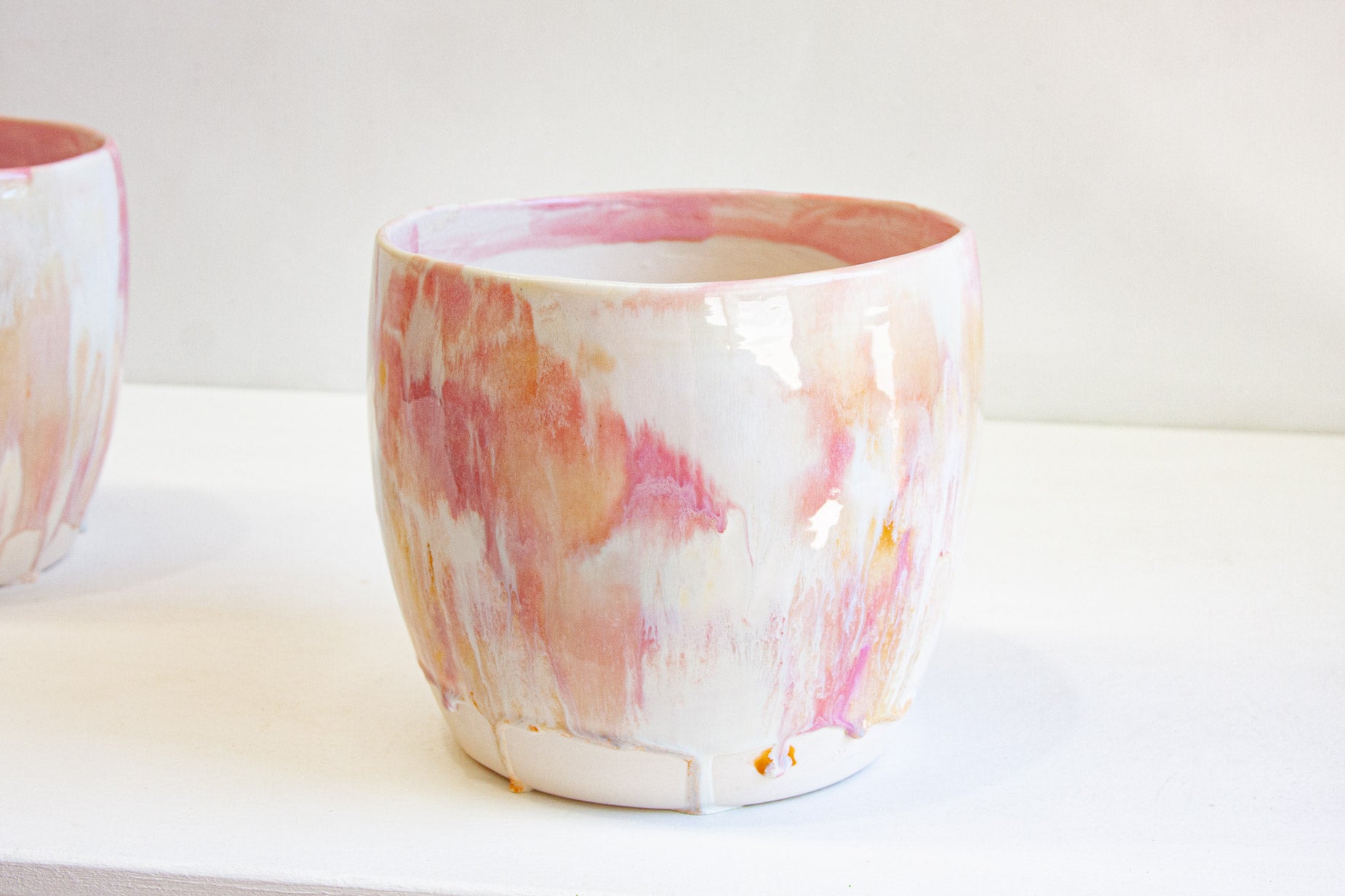 A handmade ceramic large planter glazed in pink, peach and cream, with a second planter behind it on a white surface.