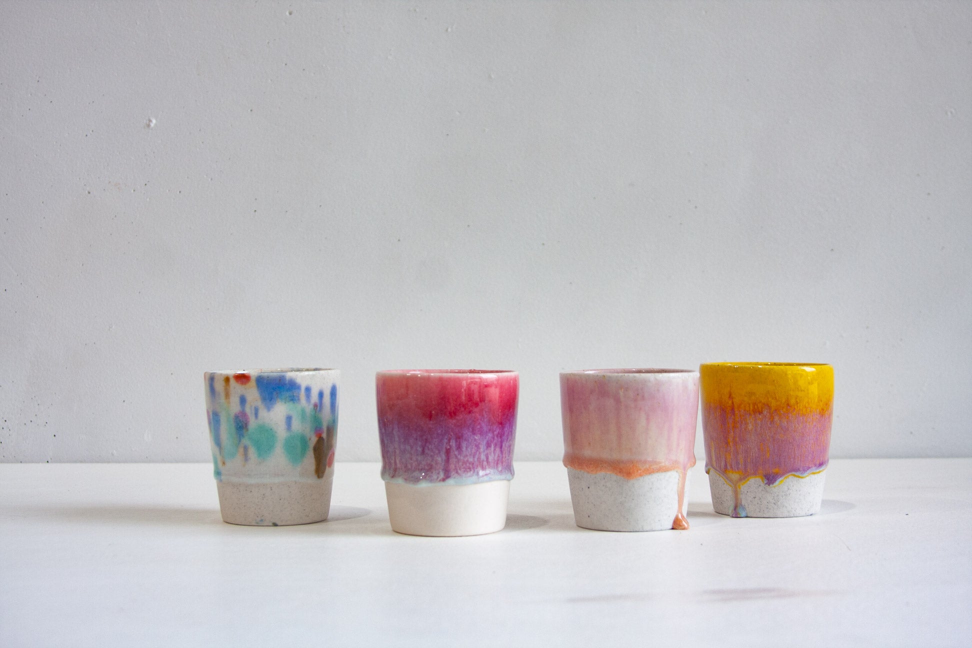 Four handmade ceramic tumblers in a row: a speckled white dabbed with blue and orange, a crimson fading into purple, a soft pink with a coral drip, and a bright yellow running into purple.