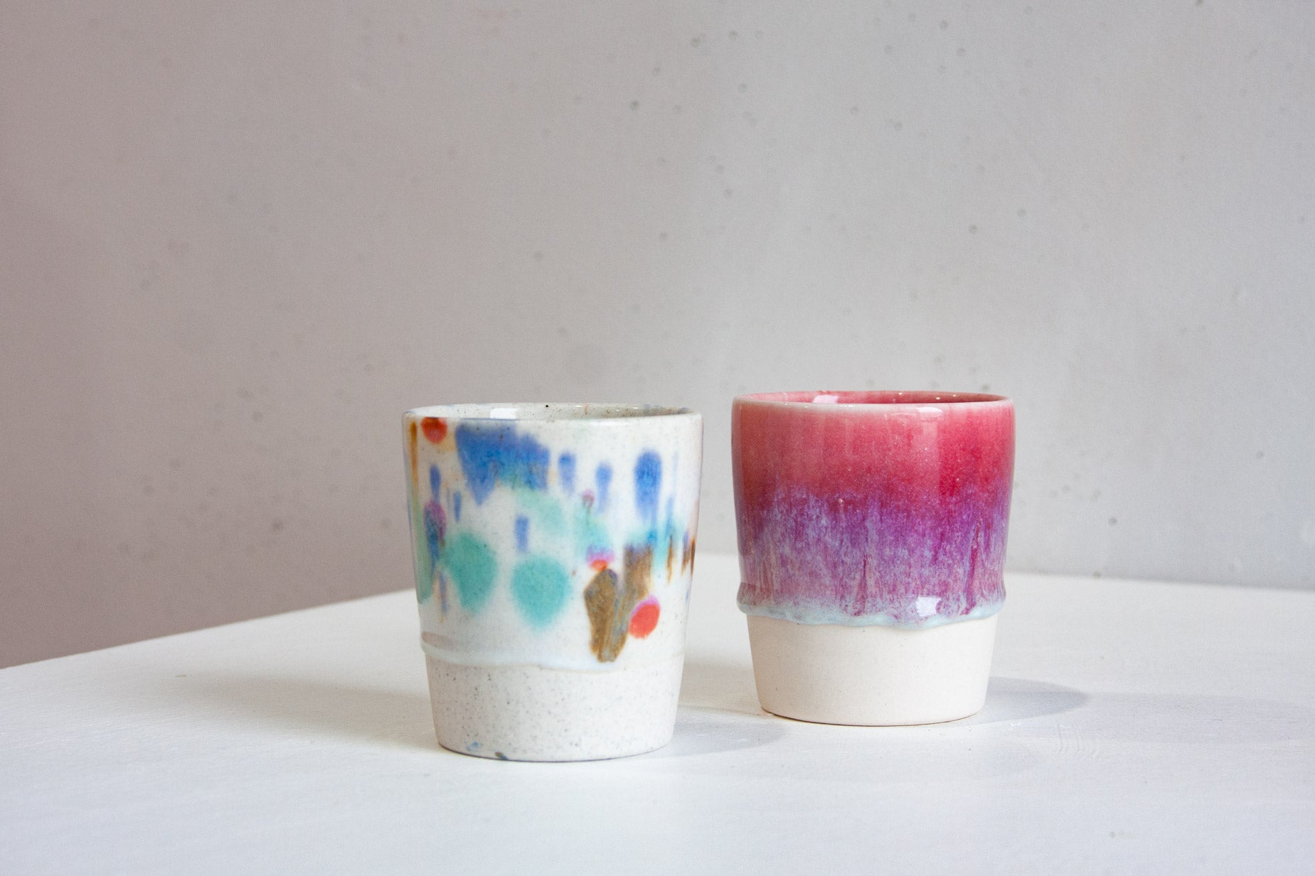 Two handmade ceramic tumblers, one a speckled white dabbed with blue, teal and orange, the other a crimson fading through purple into lilac above a bare clay base.