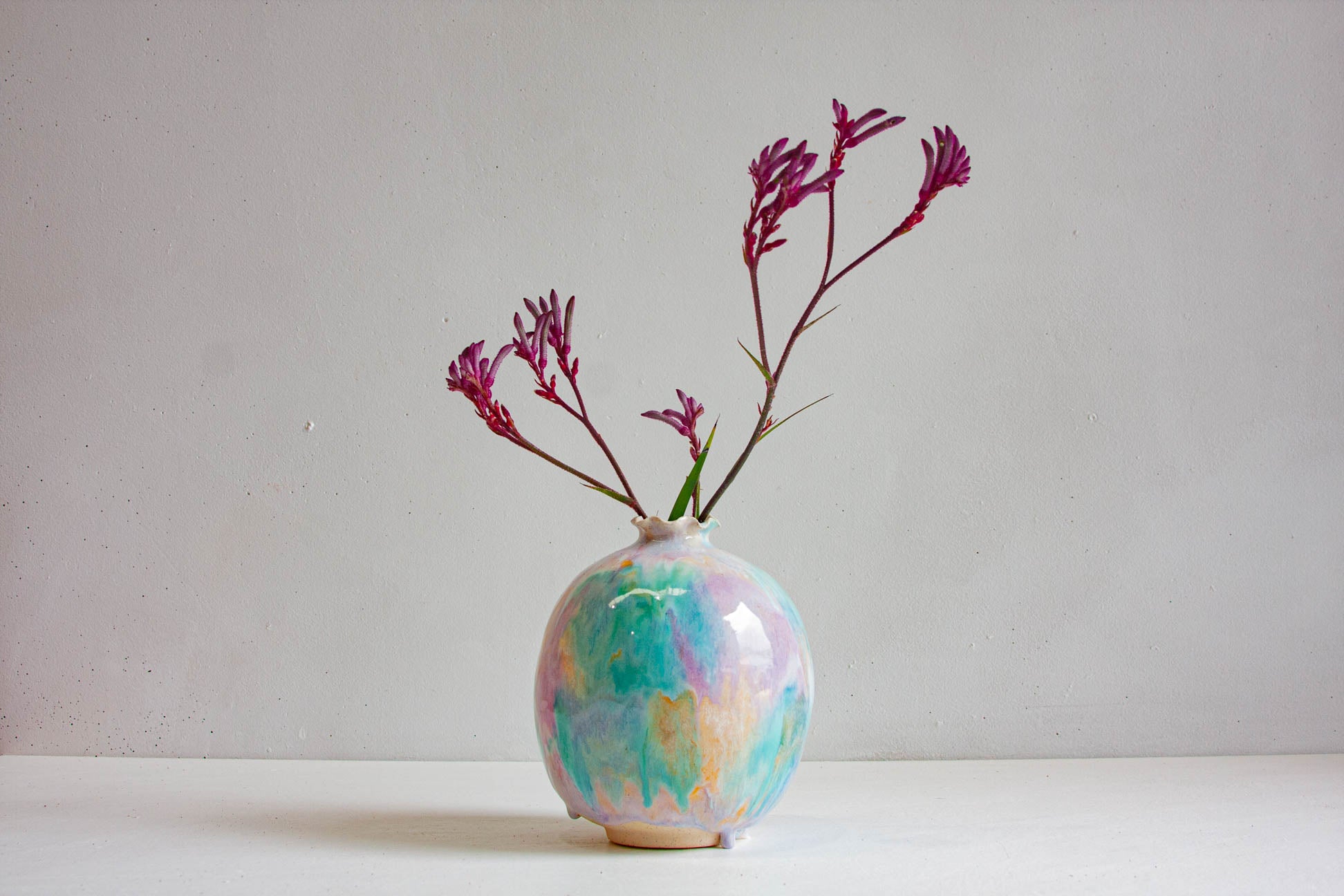 A handmade ceramic sculptural vase holding stems of kangaroo paw, the round body glazed in pale green, purple and peach.