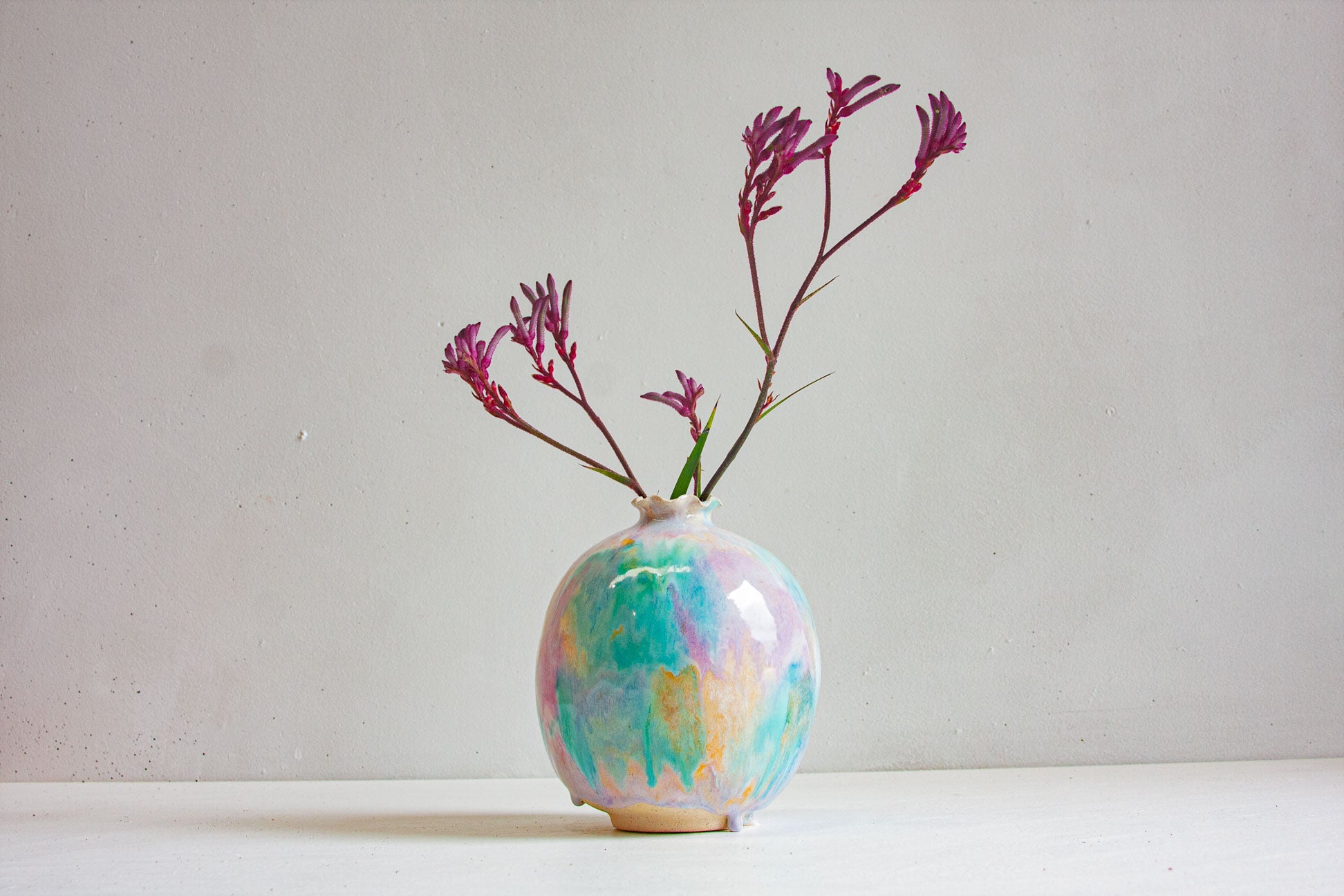 A handmade ceramic sculptural vase with a round body and small frilled neck, glazed in pale green, purple and peach, holding stems of kangaroo paw.
