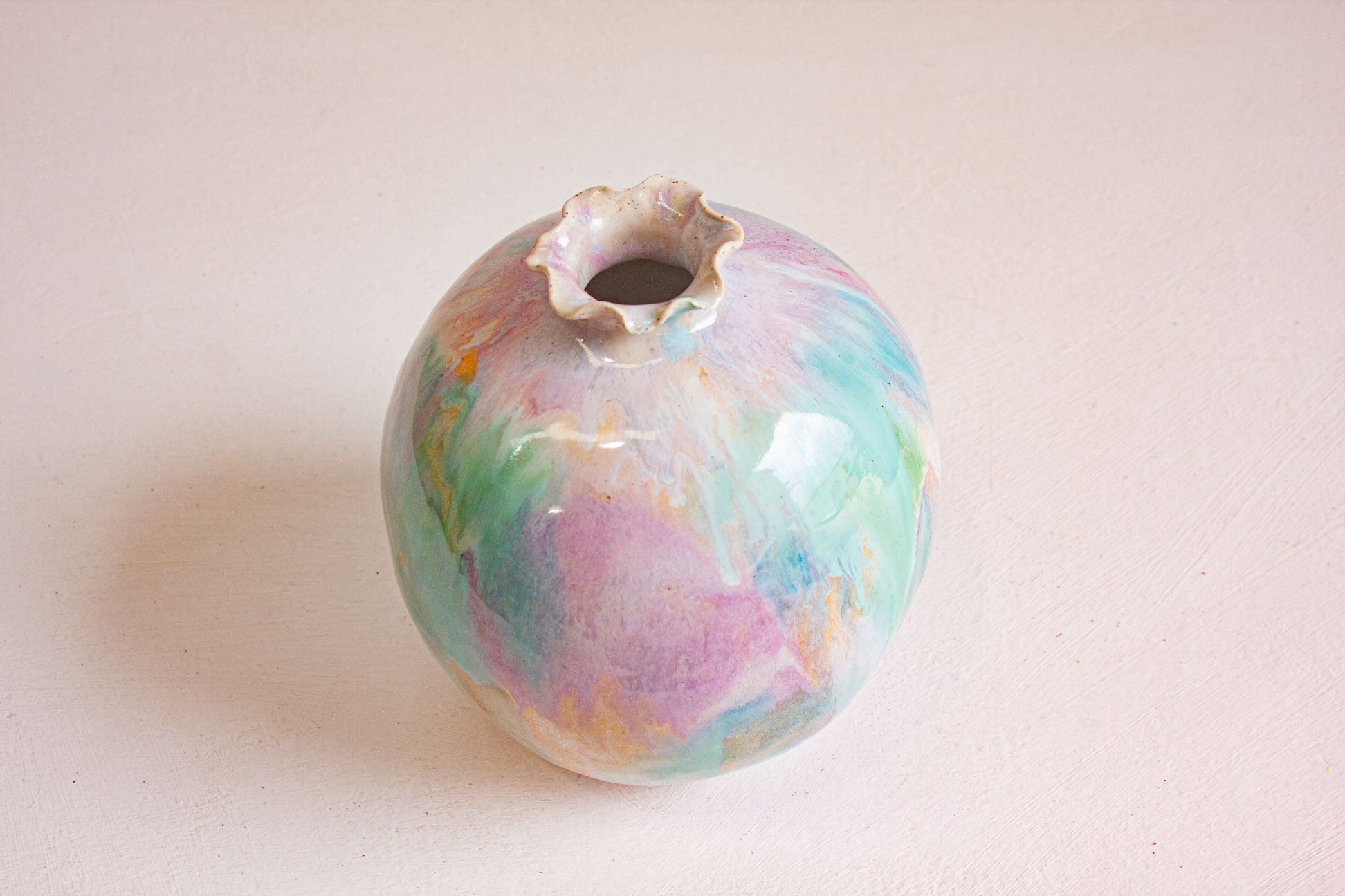 A handmade ceramic sculptural vase seen from above and to one side, the round body glazed in soft green, purple and peach.
