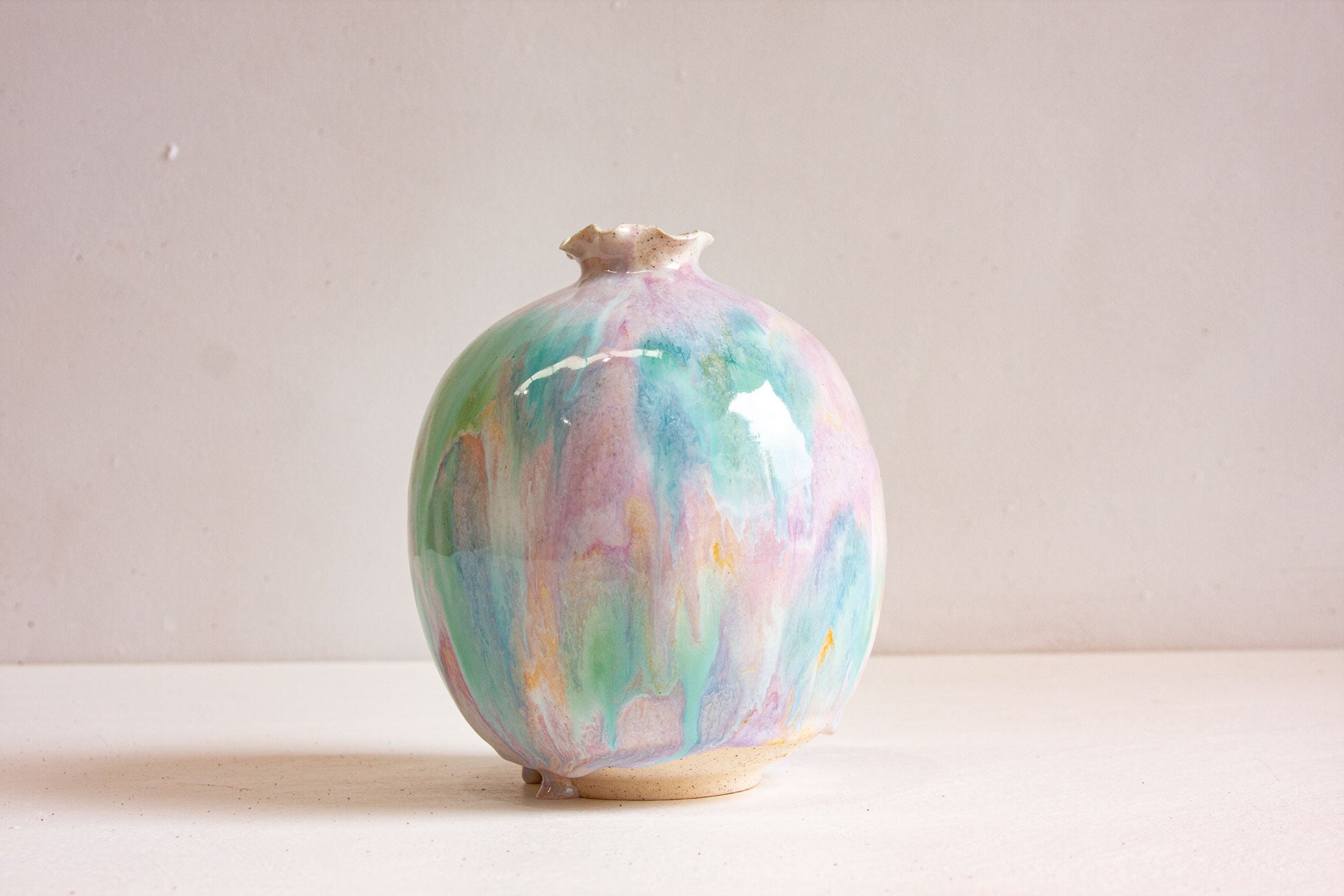 A handmade ceramic sculptural vase seen front on, pale green, purple and peach running down the round body to small feet at the base.