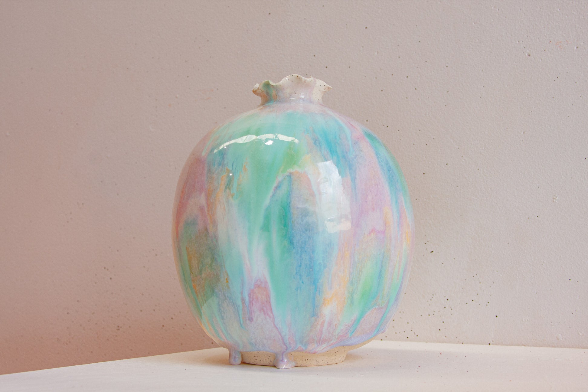 A handmade ceramic sculptural vase against a soft pink wall, the round body glazed in green, purple and peach.