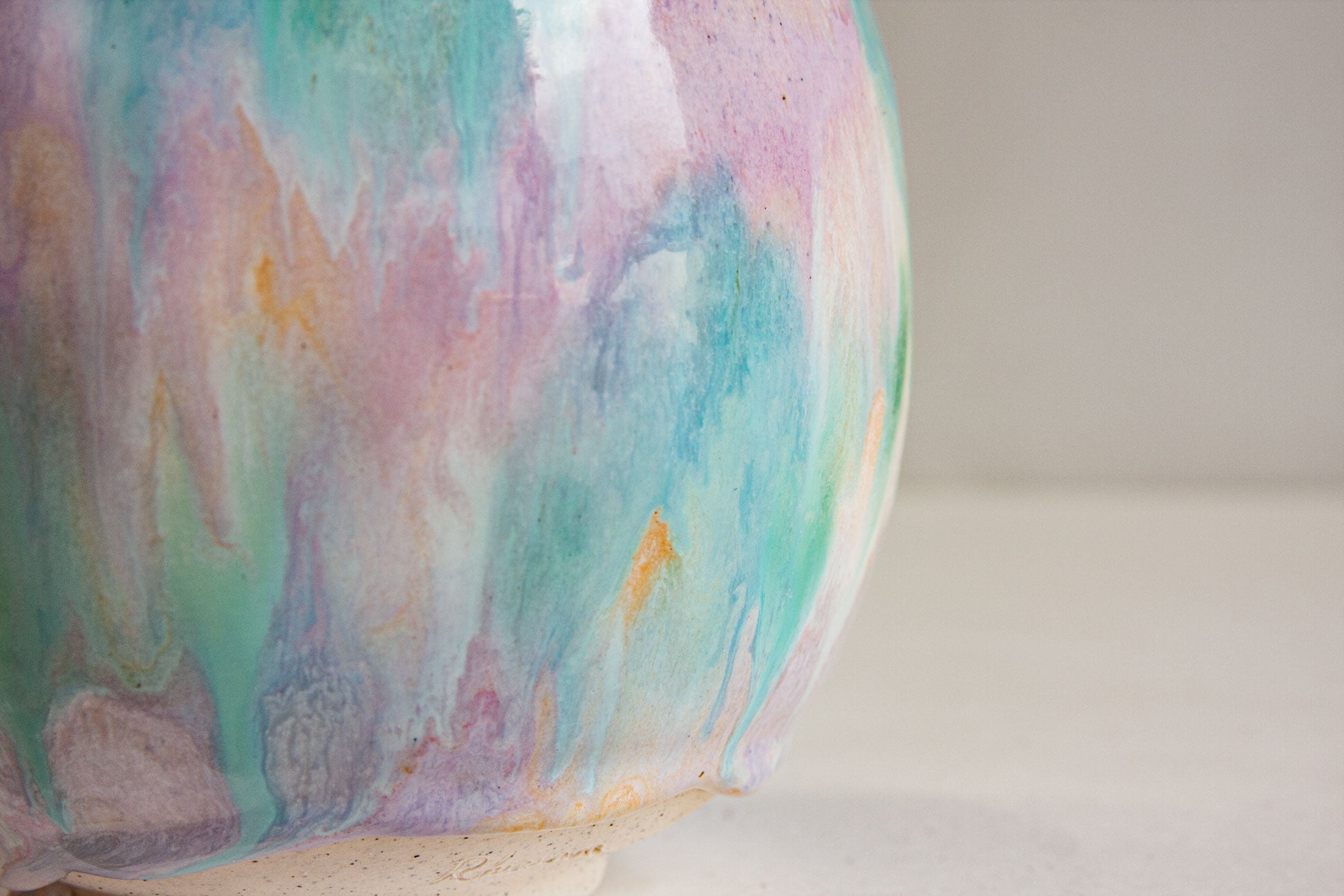 Close-up of the glaze on a handmade ceramic sculptural vase, pale green, purple and peach running in soft vertical bands over bare clay at the foot.