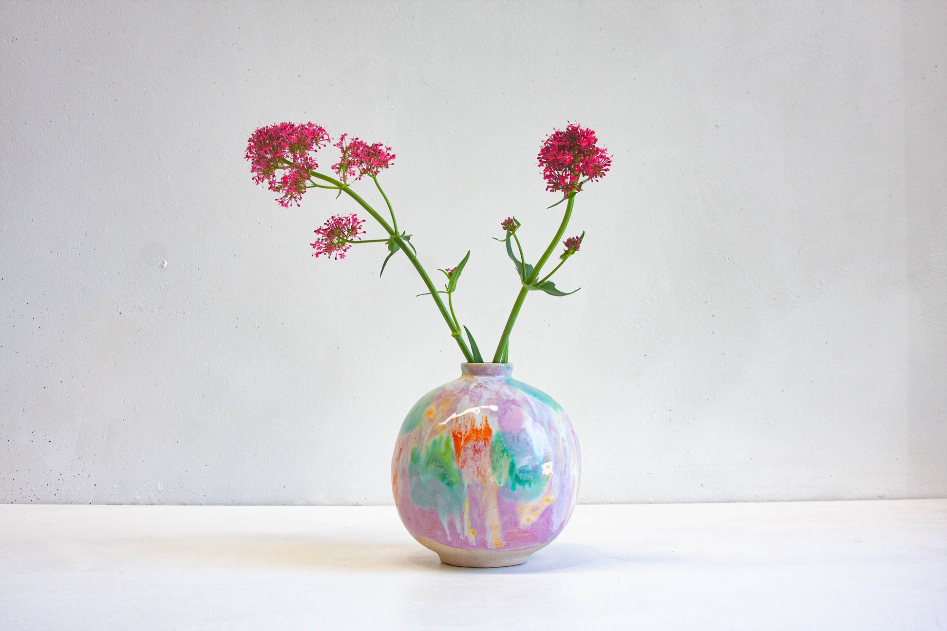 A round handmade ceramic vase glazed in purple, green, orange and yellow, holding two stems of deep pink flowers against a pale wall.