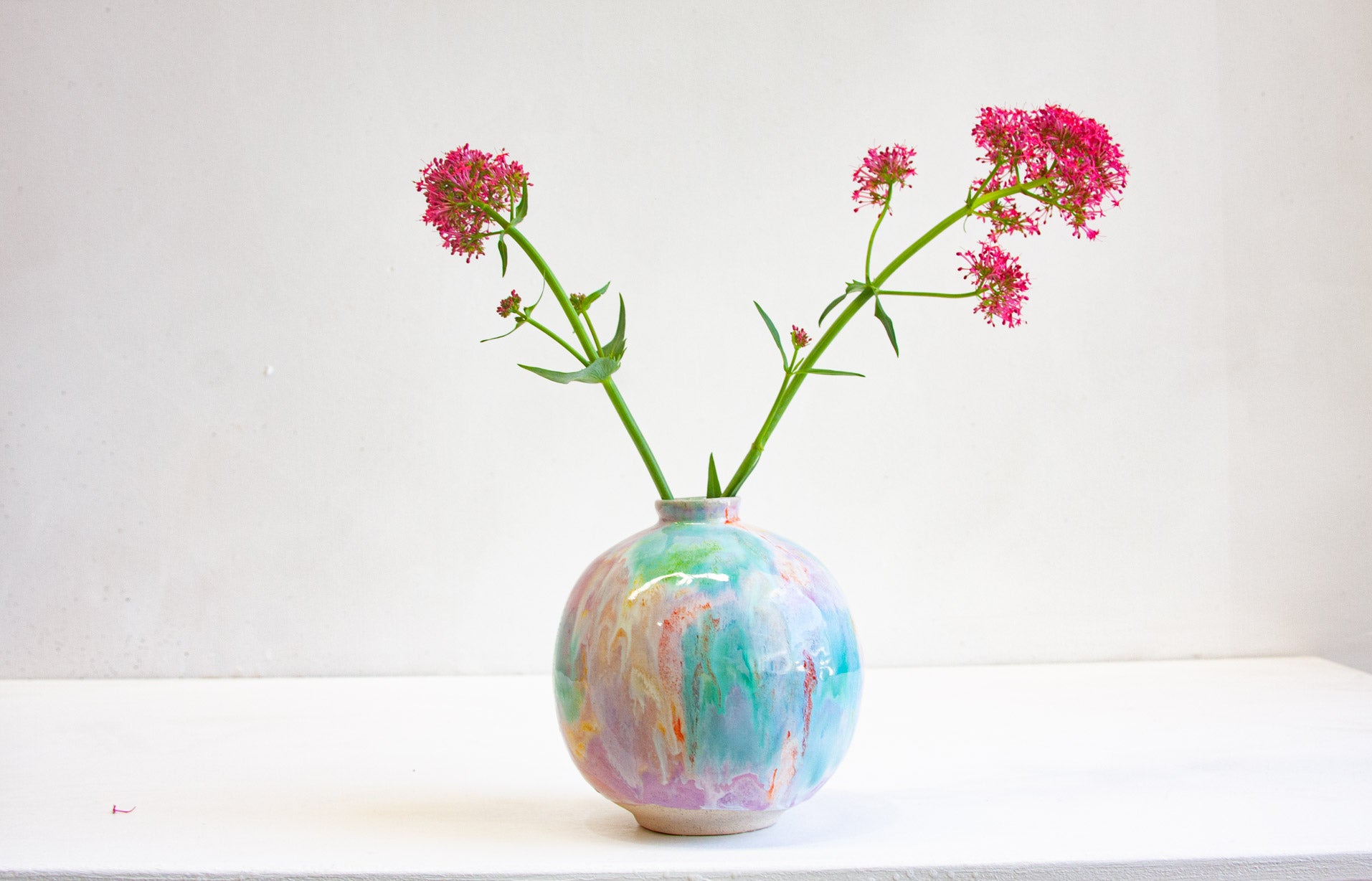A round handmade ceramic vase holding deep pink flowers, the purple, green, orange and yellow glaze washing across the full-bellied form.