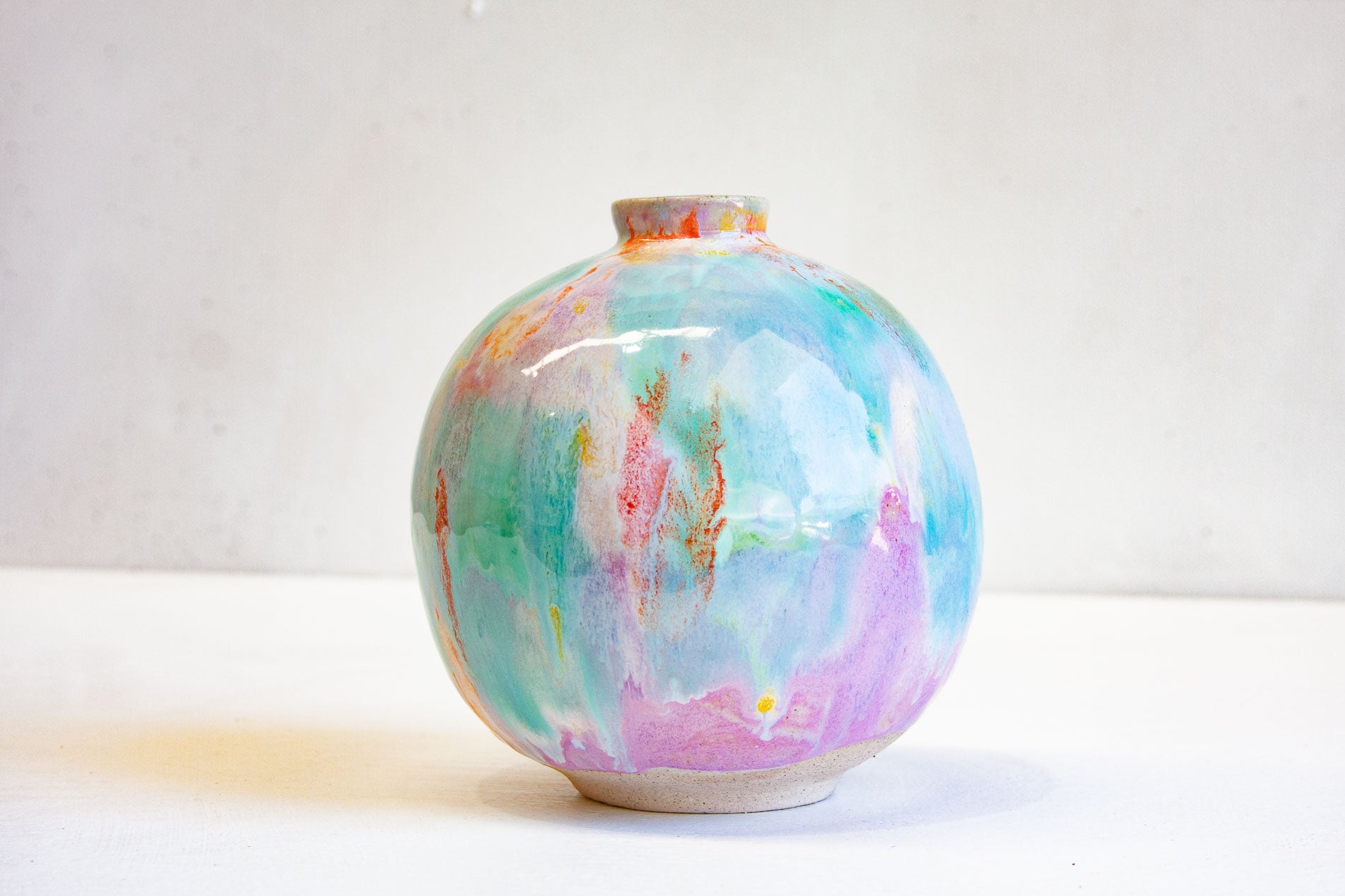 A round handmade ceramic vase photographed front on, purple, green, orange and yellow running down the body to the bare clay foot.