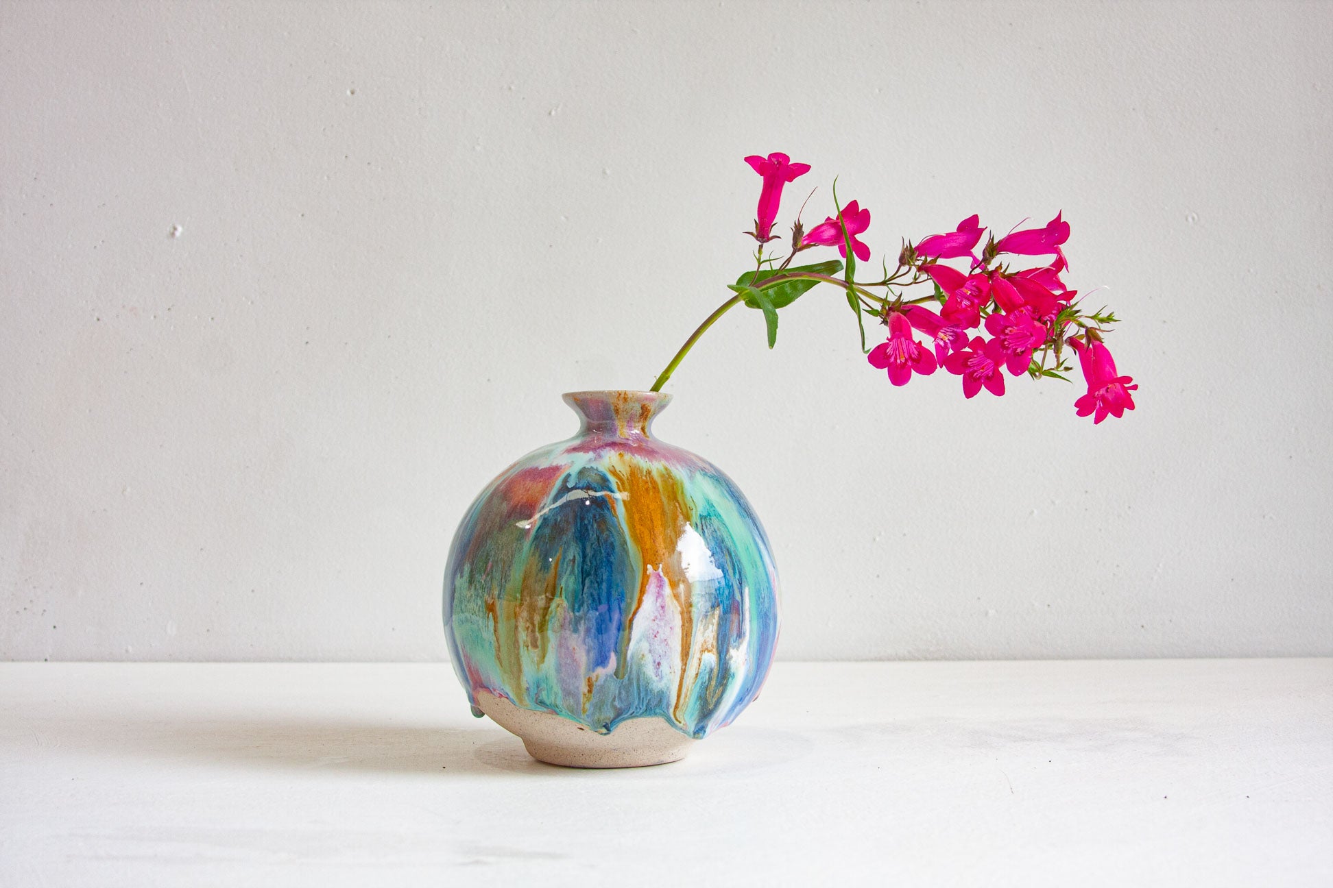 A round handmade ceramic vase glazed in blue, teal, pink, peach and ochre, holding a stem of bright pink flowers against a pale wall.