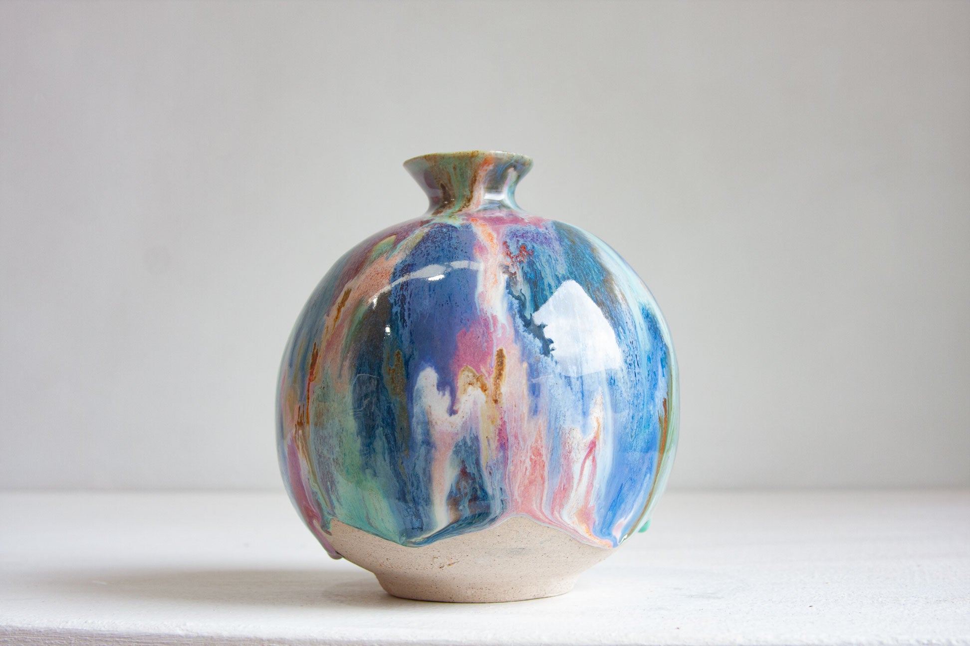 A round handmade ceramic vase photographed front on, blue and teal running down through pink and peach to the bare clay foot.