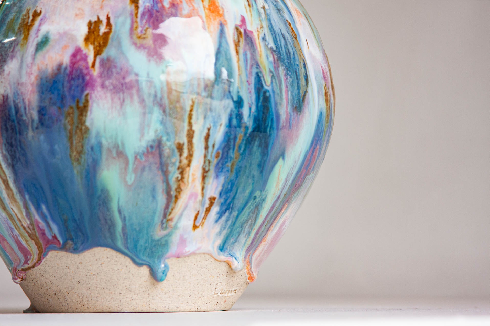 Close-up of the base of a handmade ceramic vase, the glaze stopping in drips above the bare speckled clay foot, with the maker's stamp pressed into the clay.
