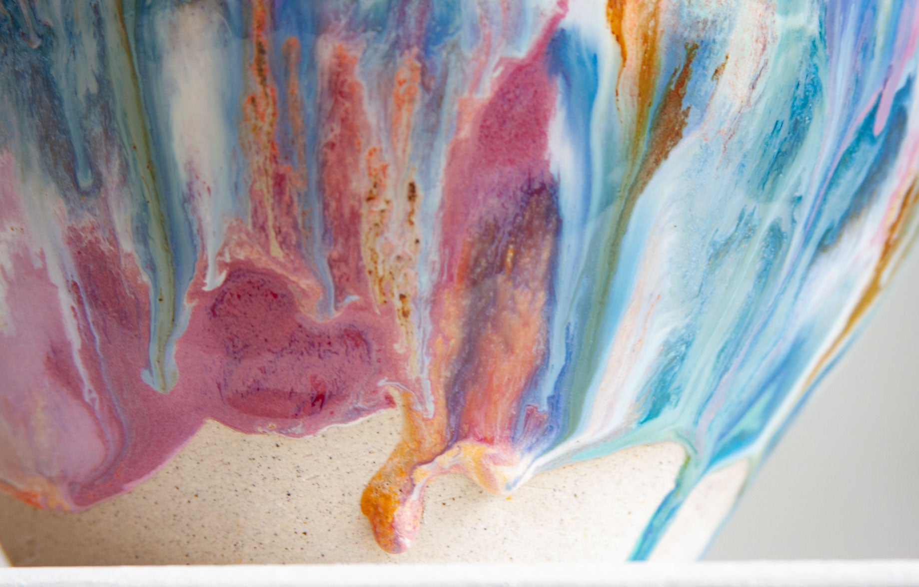 Extreme close-up of the glaze meeting the bare clay, pink, blue and ochre running together into a single drip.