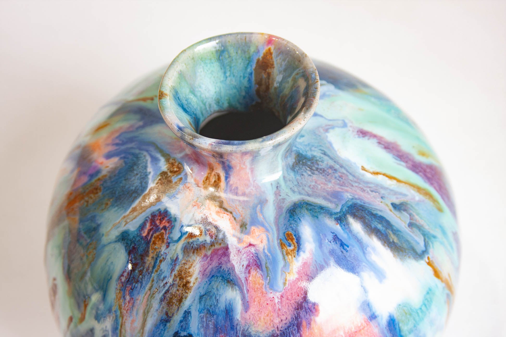 Close-up of the flared neck of a handmade ceramic vase, the blue, pink and ochre glaze pooling around the opening.