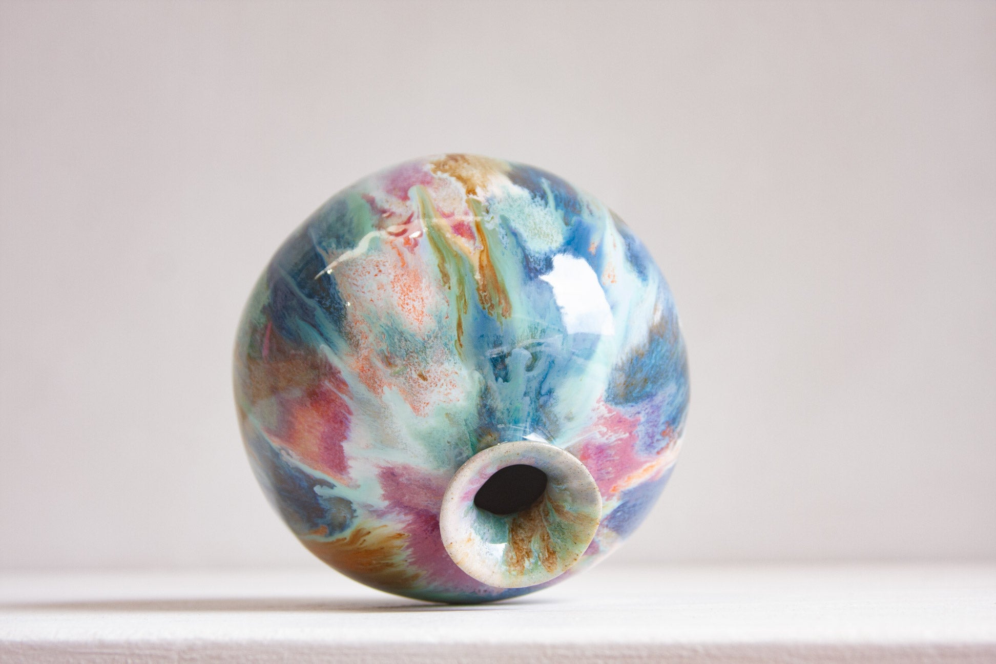 A round handmade ceramic vase resting on its side, looking into the flared neck, the glaze marbled in blue, teal, pink and ochre.