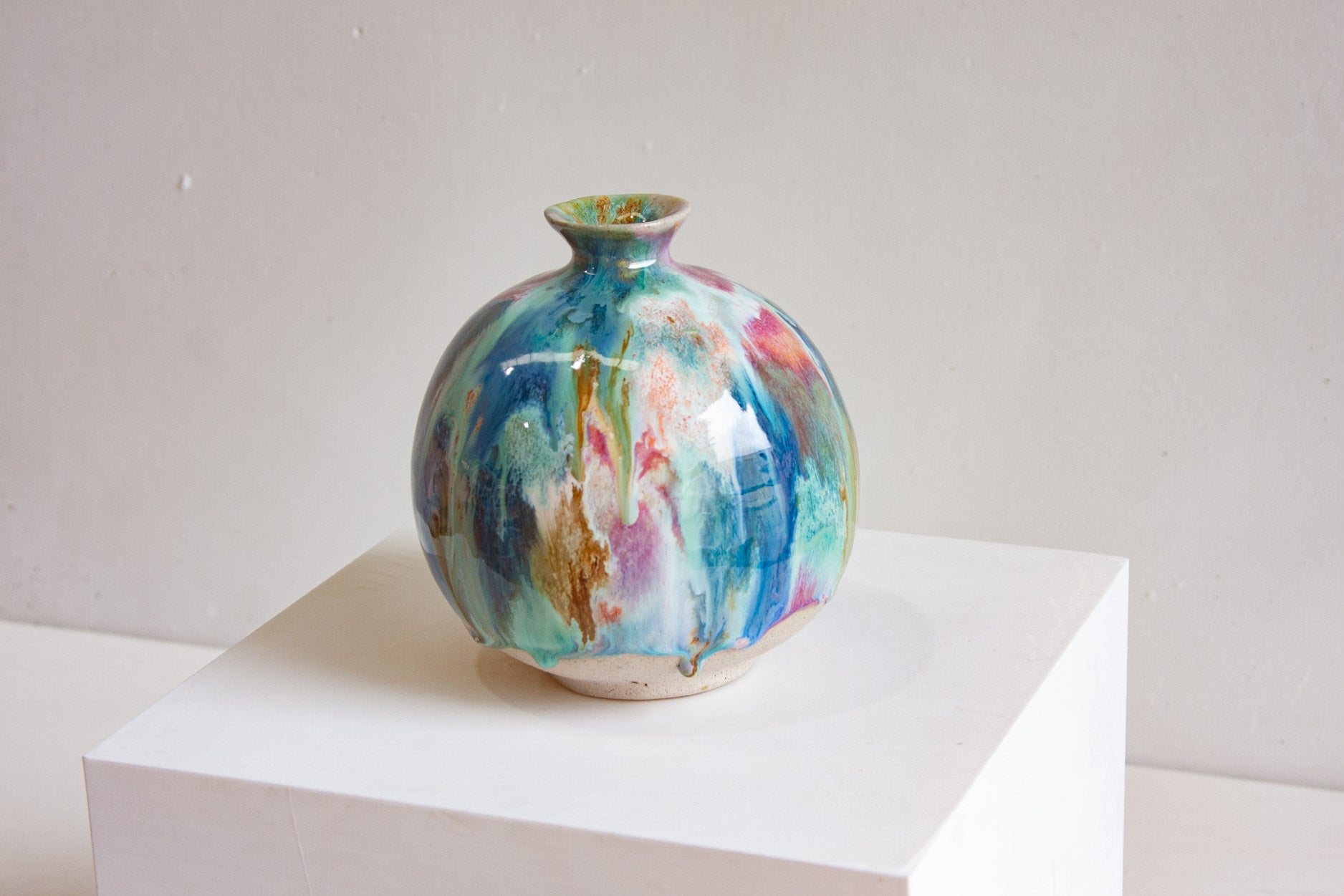A round handmade ceramic vase on a white plinth, the blue, teal, pink and ochre glaze falling in vertical streaks over the globe form.