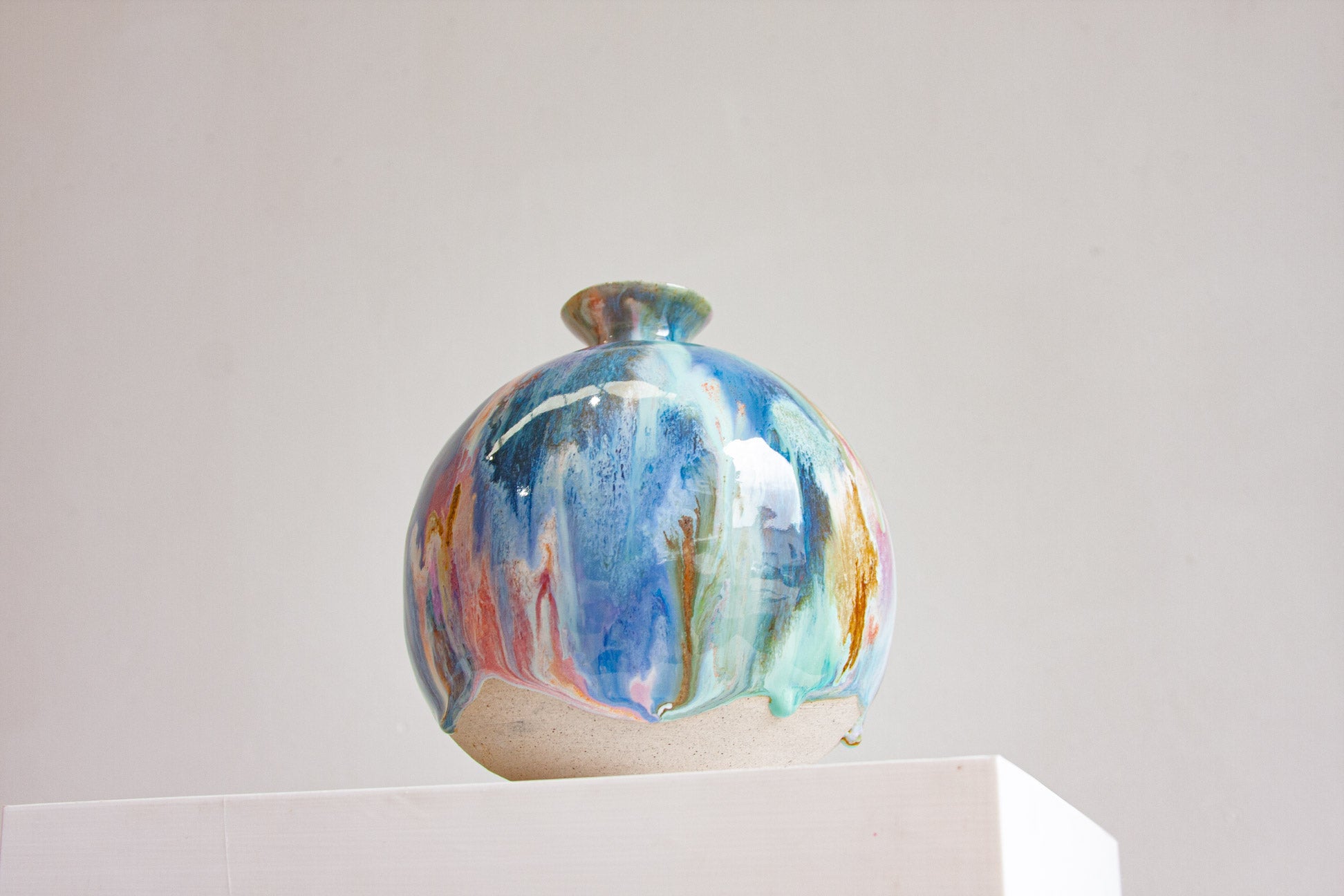 A round handmade ceramic vase seen on a plinth, the glaze running in blue, green, peach and ochre and stopping in drips above the bare clay.