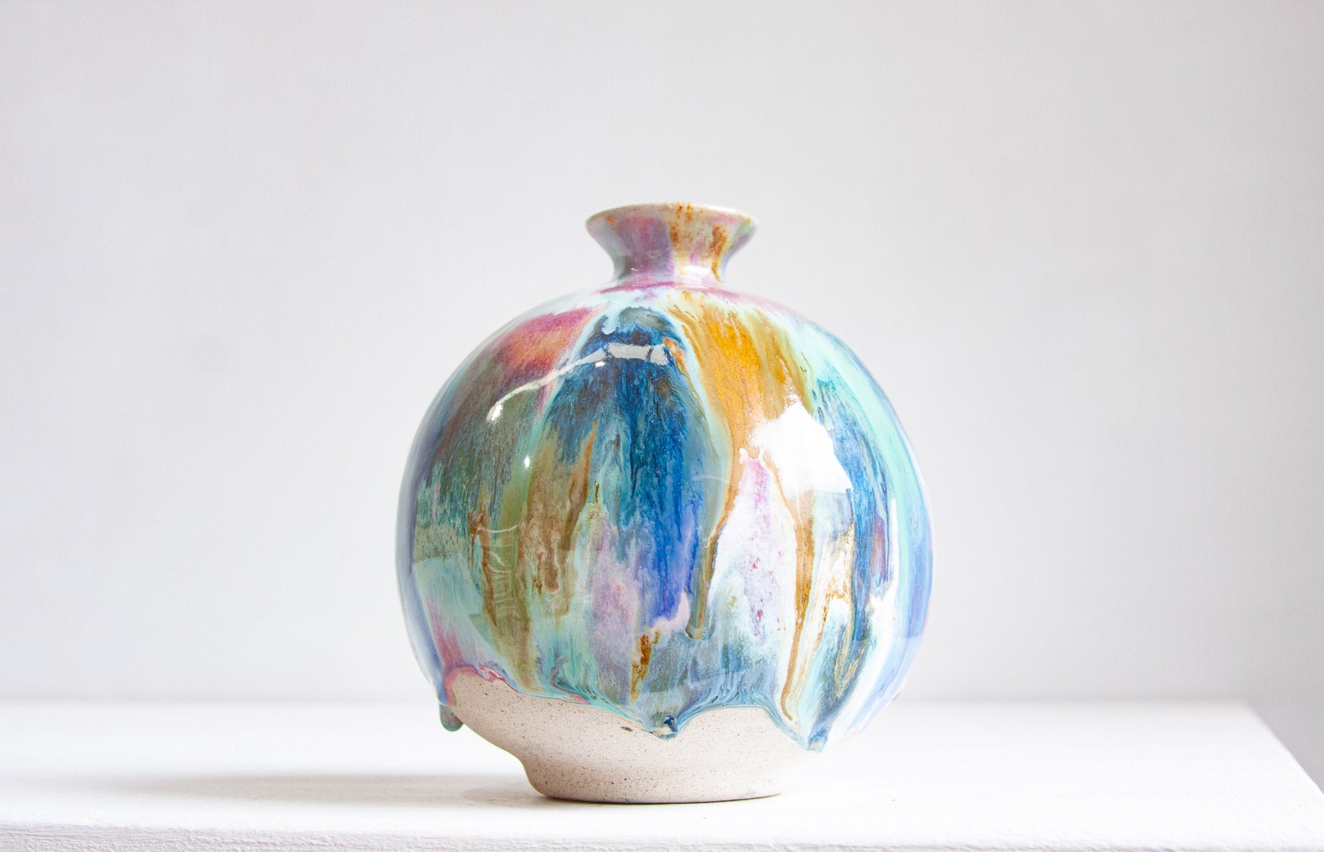 A round handmade ceramic vase front on, blue, teal, pink, peach and ochre drifting down the body to a bare speckled clay foot.