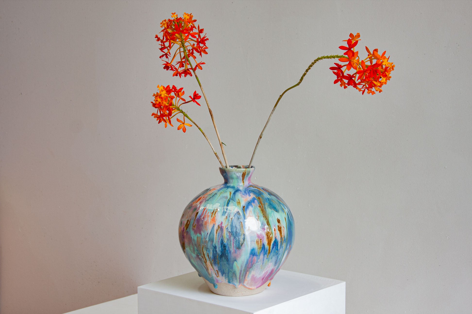 A large round handmade ceramic vase on a white plinth holding orange flowers, the blue and teal glaze running down the globe form.