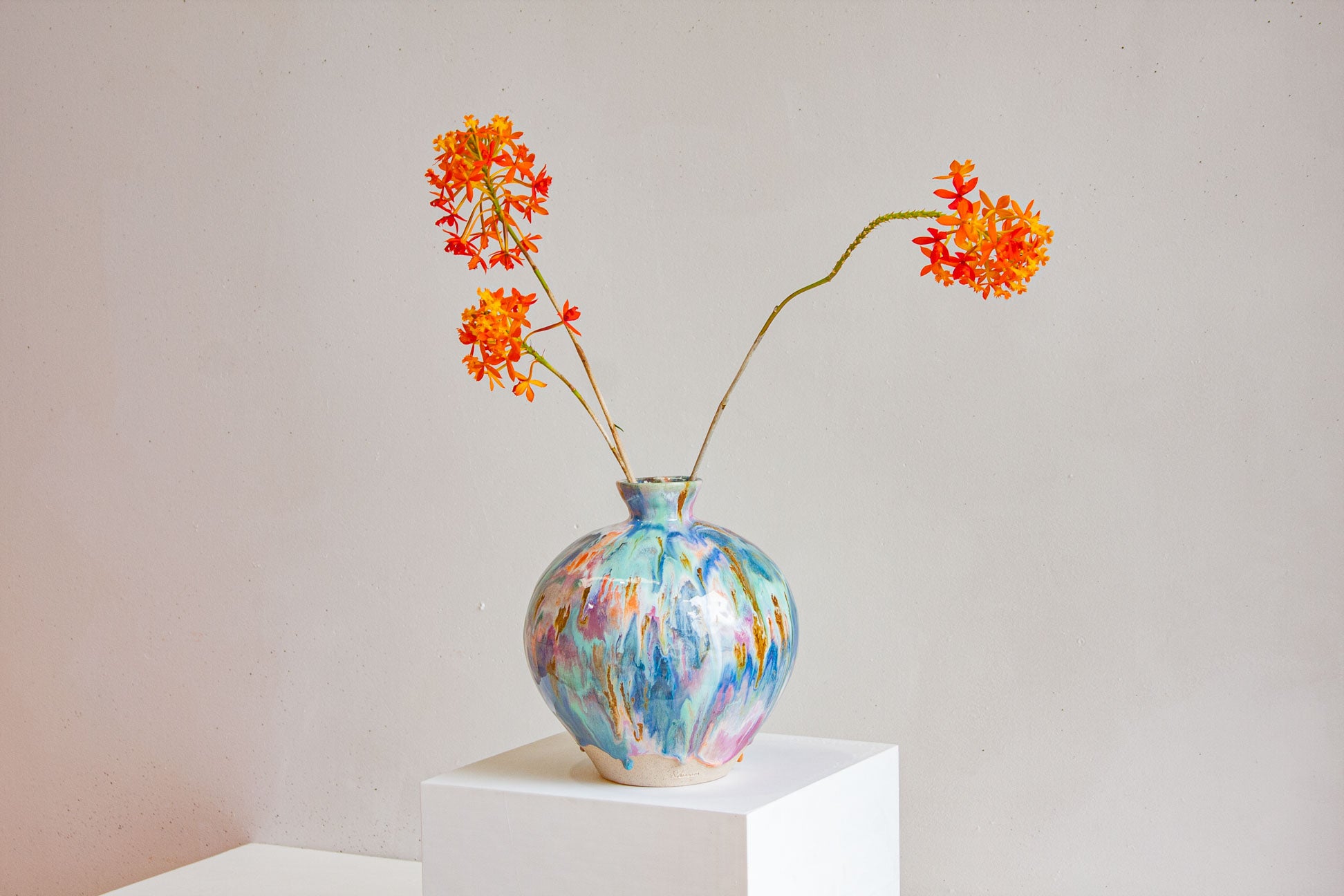 A large round handmade ceramic vase on a white plinth, glazed in blue, teal, pink and ochre, holding two stems of orange flowers.