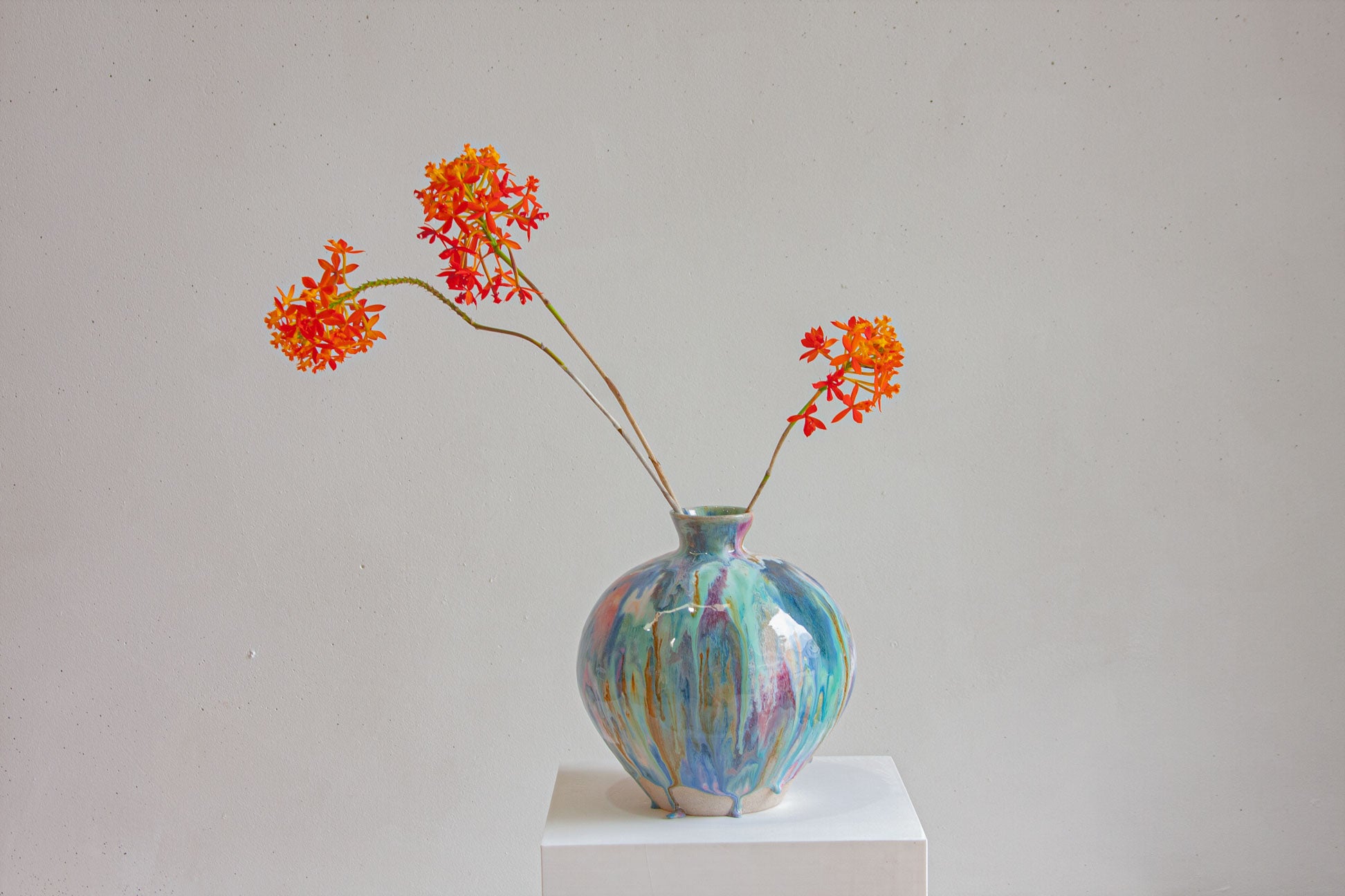 A large round handmade ceramic vase on a white plinth with orange flowers arching out of it, against a plain pale wall.