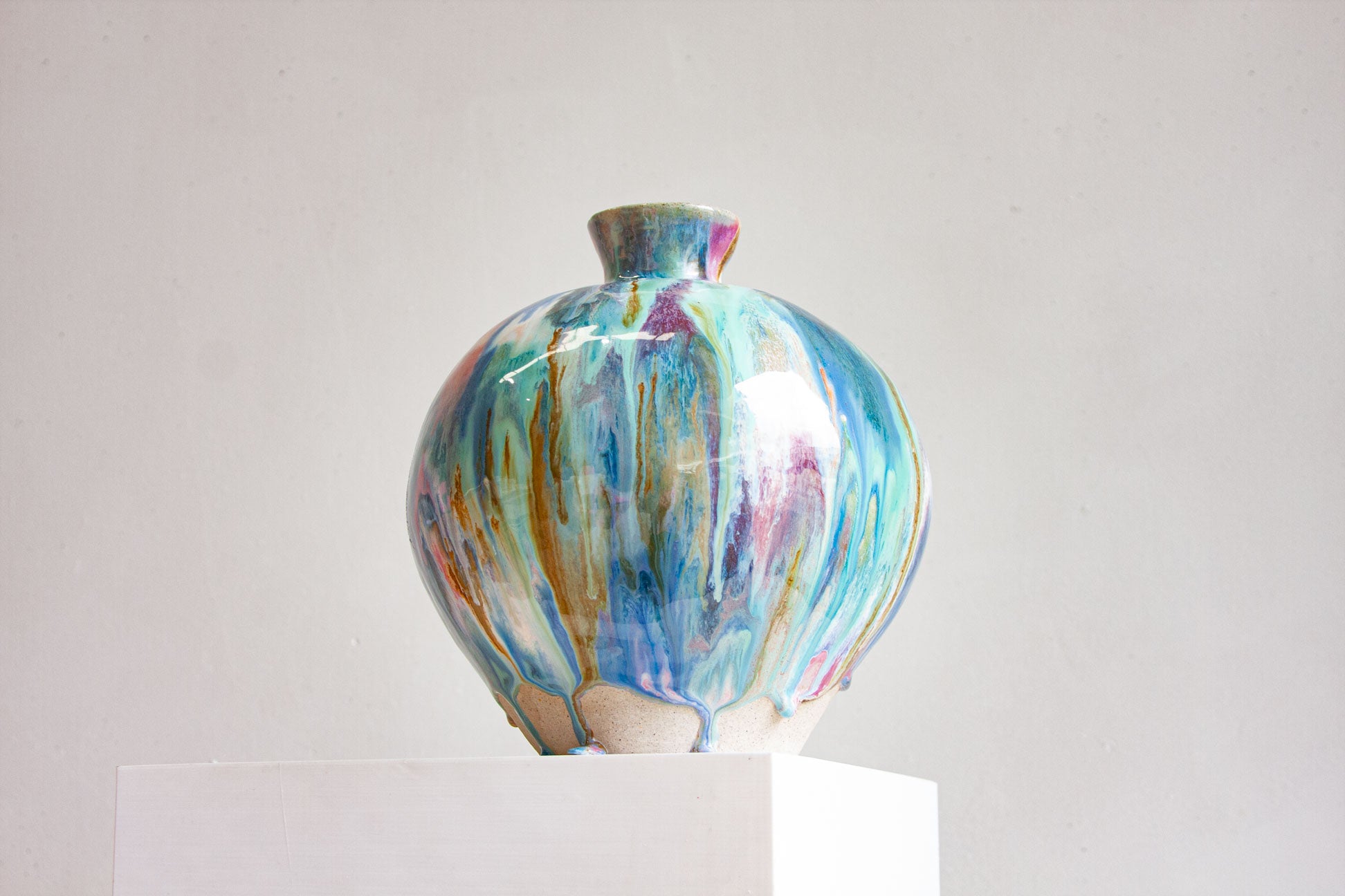 A large round handmade ceramic vase on a white plinth, front on, the glaze falling in blue, teal, pink and ochre to a bare speckled clay foot.