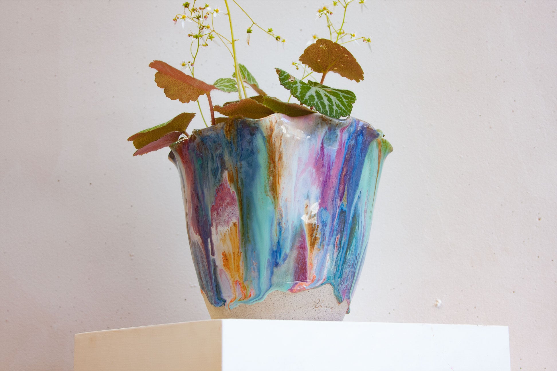 A handmade ceramic frill vessel on a plinth holding a trailing plant, the glaze running in blue, teal, purple and orange down to a bare white clay foot.