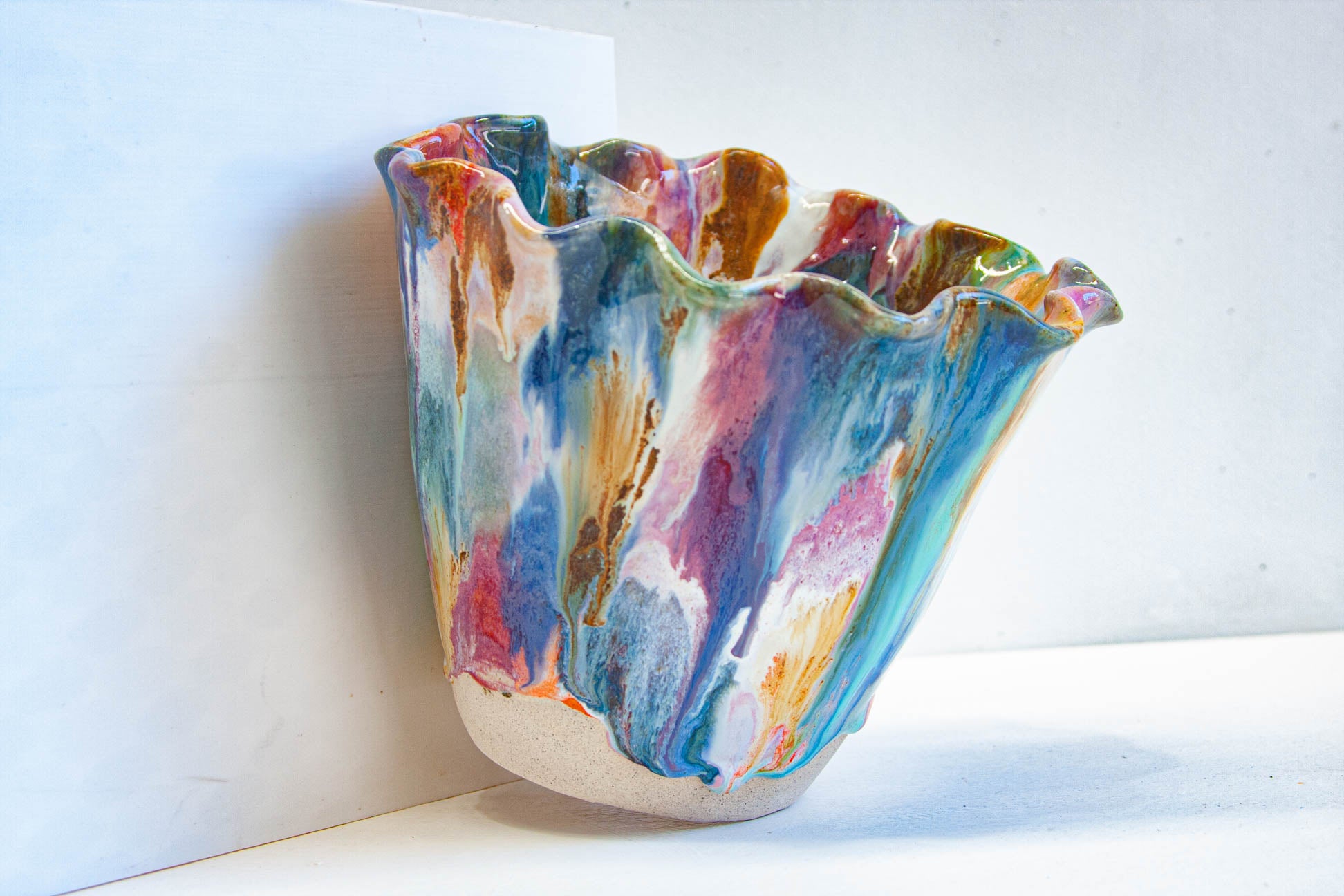 A handmade ceramic frill vessel seen from the side, blue, teal, purple, pink and orange running down the flared sides to a bare white clay foot.