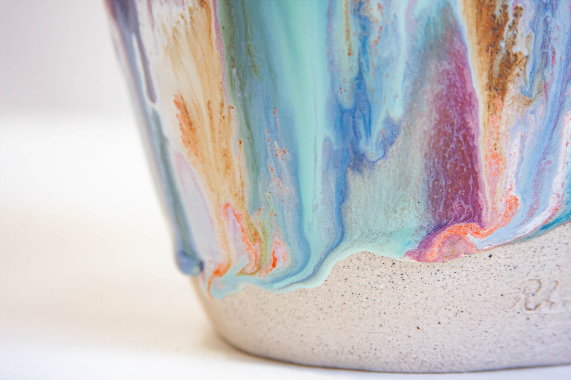 Close-up of the lower half of a handmade ceramic frill vessel, the glaze running in vertical streaks of blue, teal, purple and orange above a bare white clay foot.