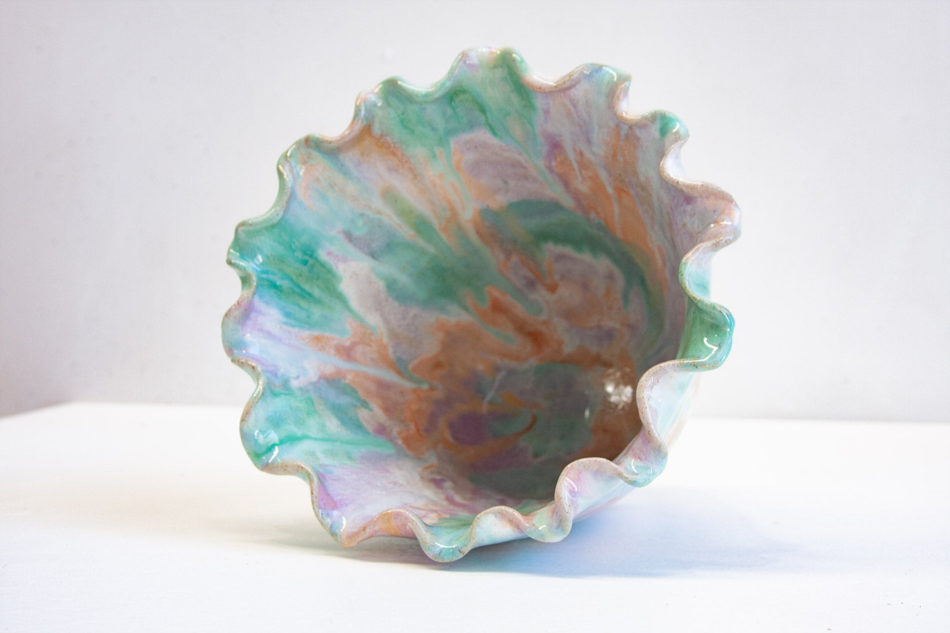 A handmade ceramic frill vessel seen from above and to one side, the frilled rim curving around an interior glazed in purple, peach and green.